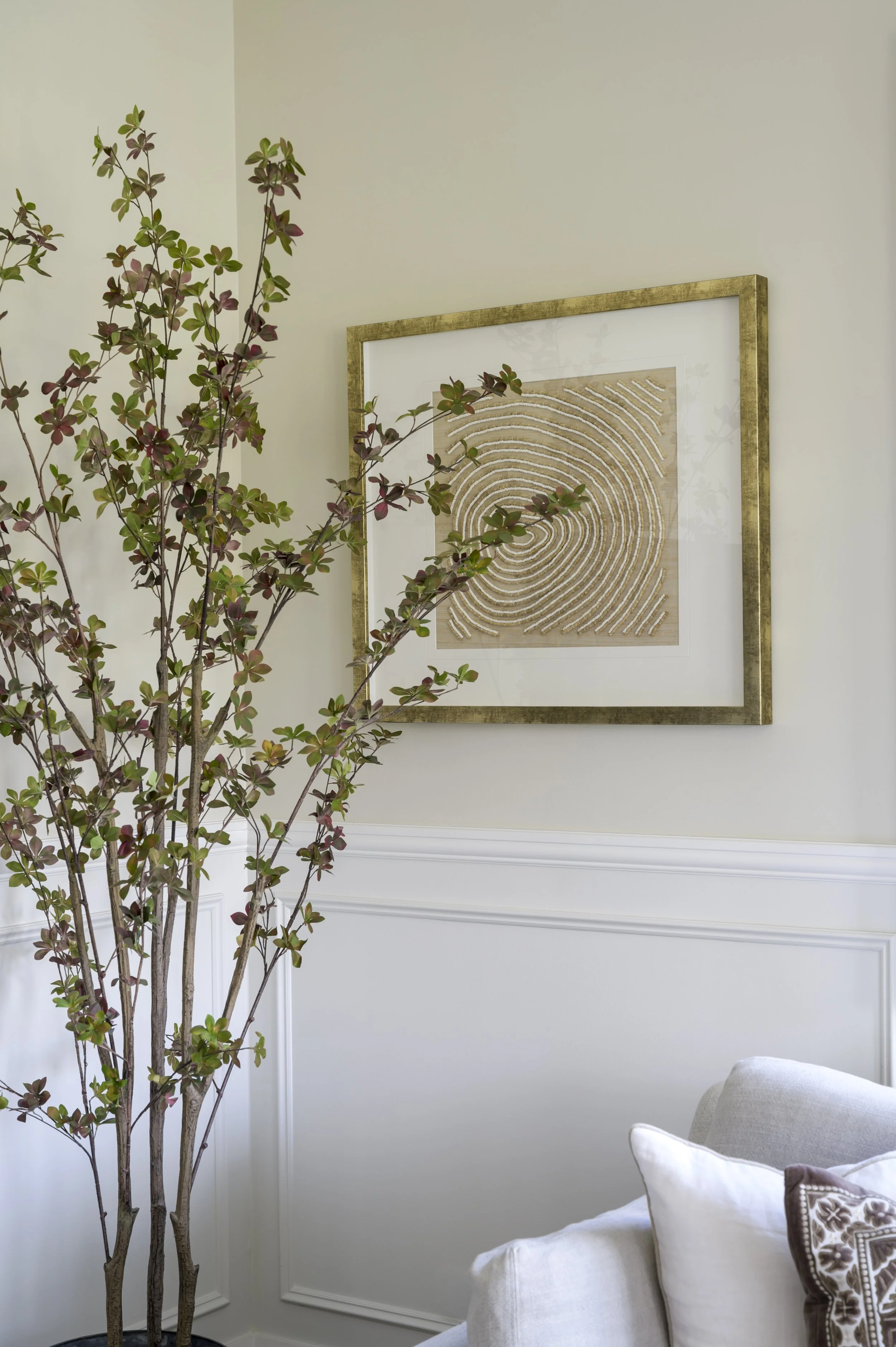 Interior corner with white wainscoting, a framed beige and white abstract art piece on the wall, a tall leafy potted plant, and a white sofa with decorative pillows.
