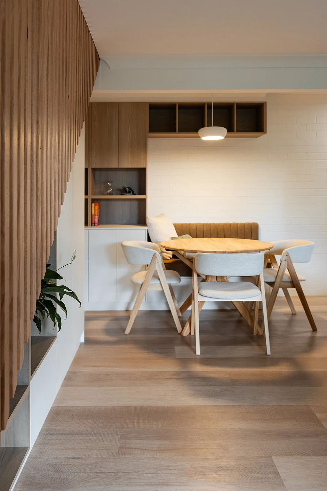 Dining area with a round wooden table, four beige upholstered chairs, a brown upholstered bench with pillows, a white cabinet, open wood shelving, and a hanging light fixture, in a modern room with wood and white walls and wood flooring.