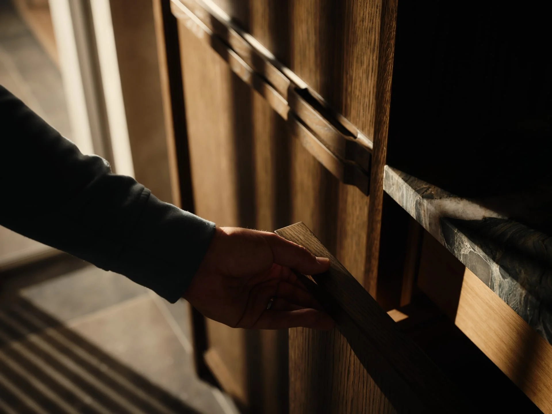 A person reaching into a wooden cabinet with a hand. The cabinet has multiple drawers and a marble countertop. The person is wearing a dark jacket.