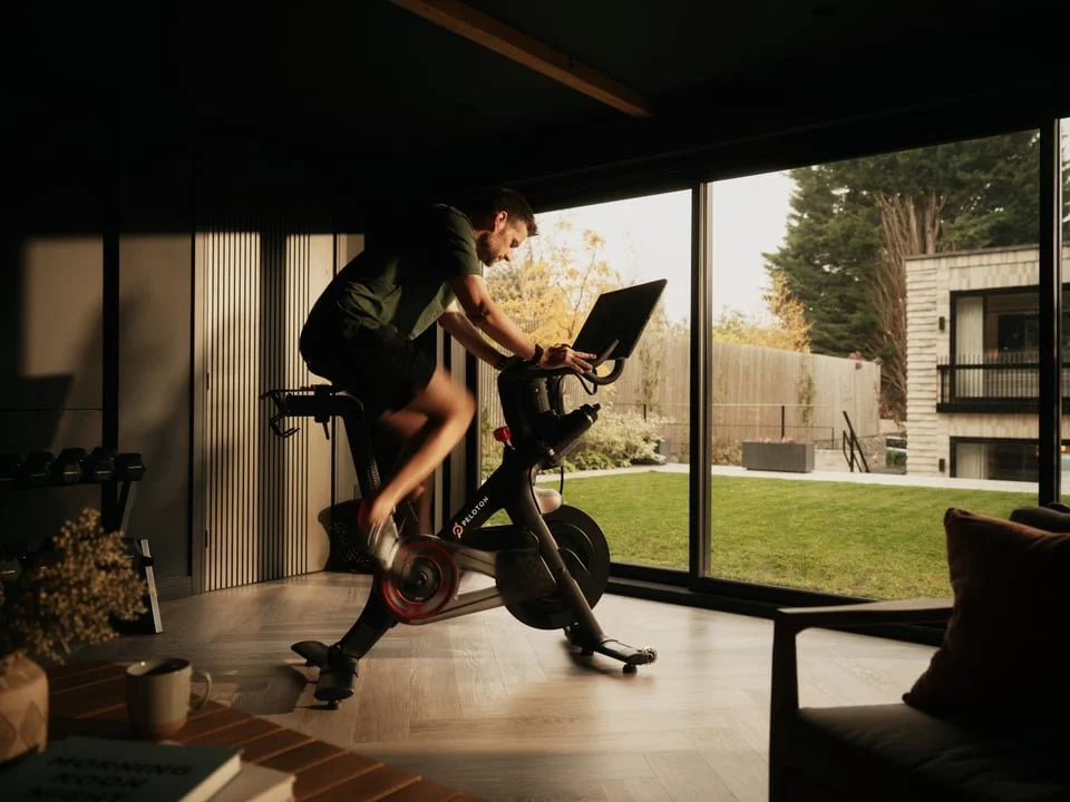 Person riding a stationary exercise bike inside a room with large glass windows showing an outdoor yard.