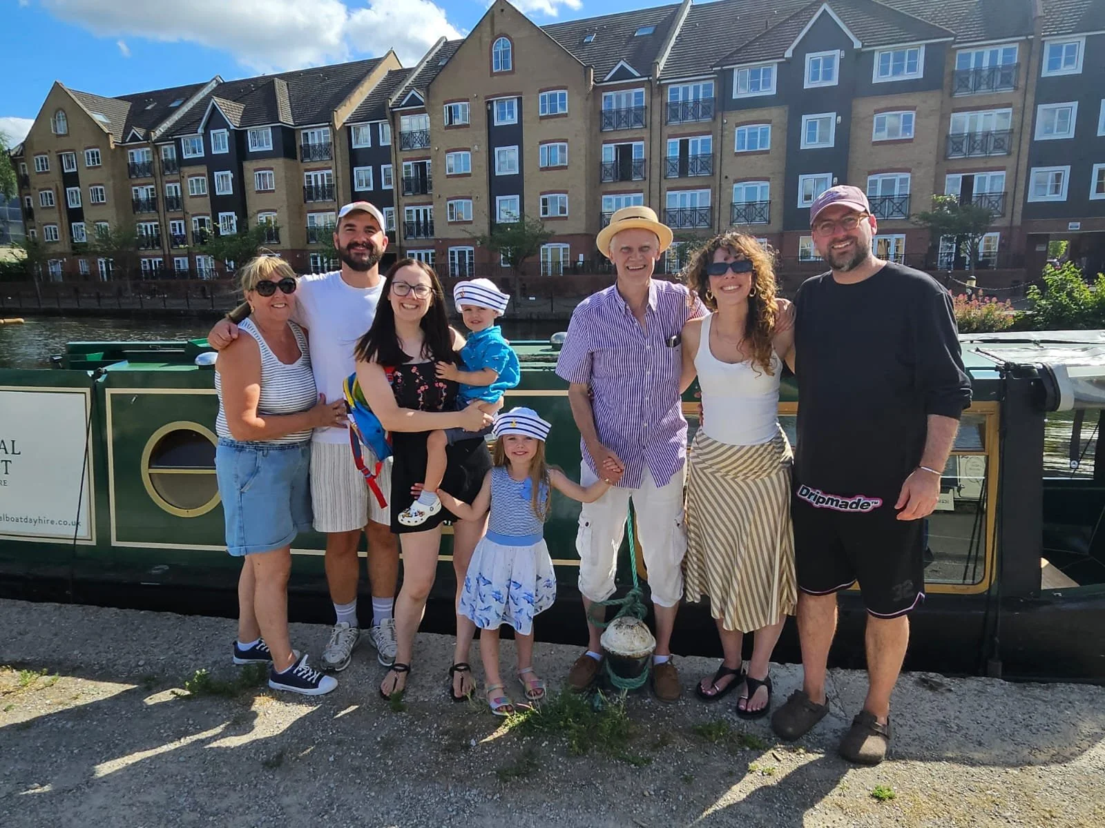 A group of eight people, including children, standing together outdoors near a canal with Tilly the narrowboat behind them. They are smiling and dressed casually, with a large residential building in the background.