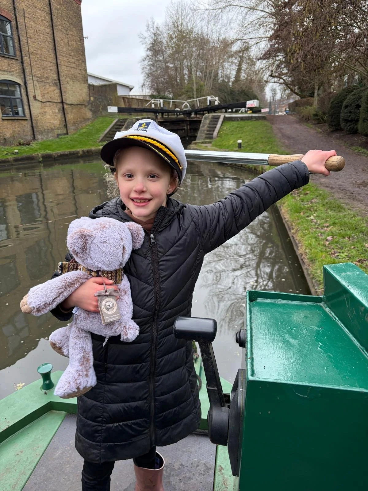 A smiling young girl wearing a captain's hat, holding a plush teddy bear with a badge, and standing on Tilly the canal boat. She is pointing with her arm extended to the right, with a canal lock tower and trees in the background.