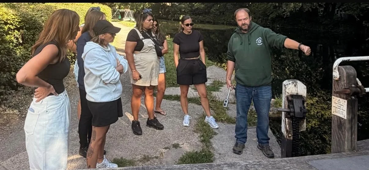A group of six women and one man gathered outdoors by a small waterway, with the man explaining how to open the locks using a windlass, while the women listen attentively.