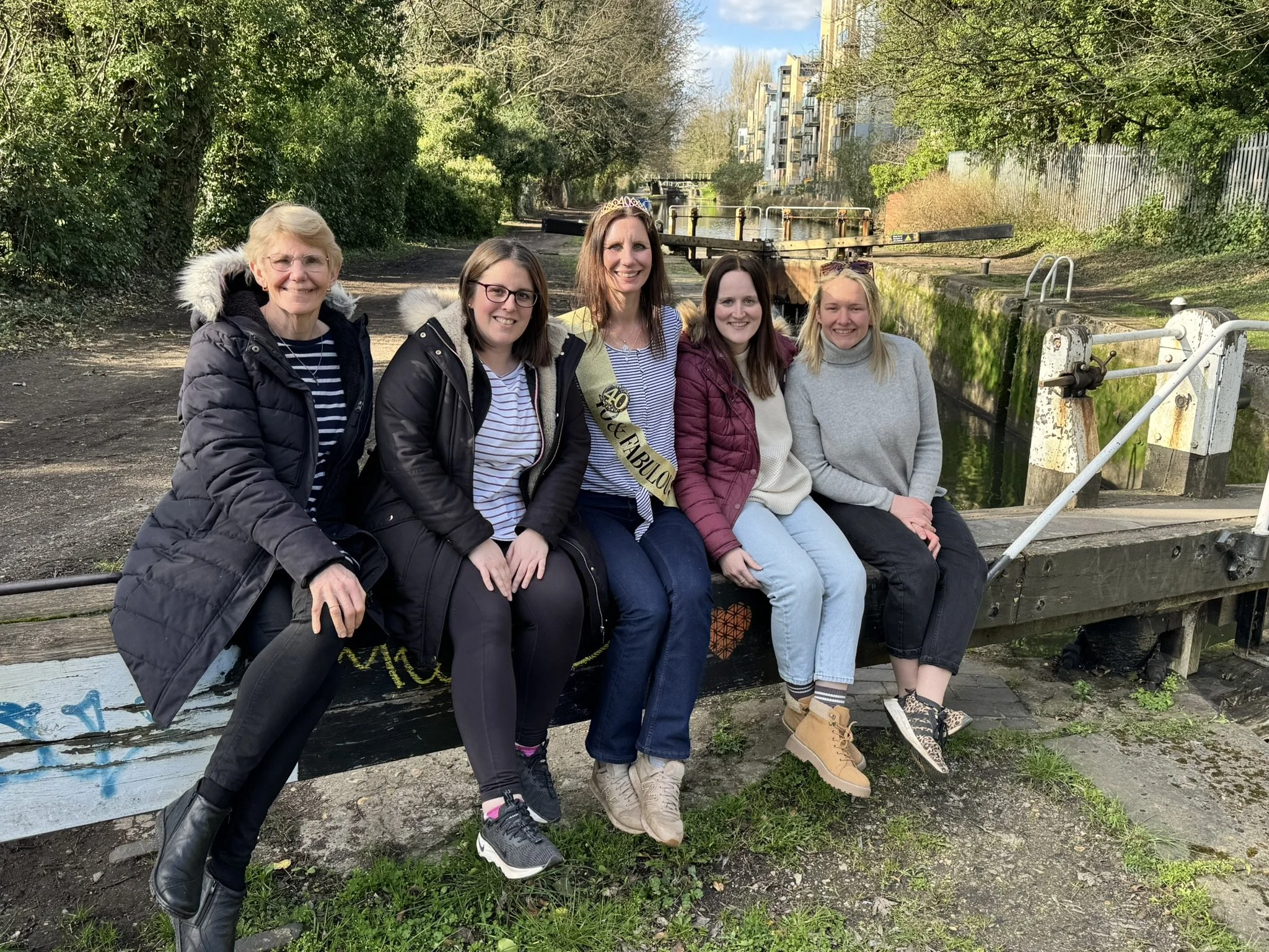 Group of women sitting on a park bench celebrating a birthday on Tilly on the Grand Union Canal in Apsley with trees, waterway, and buildings in the background.