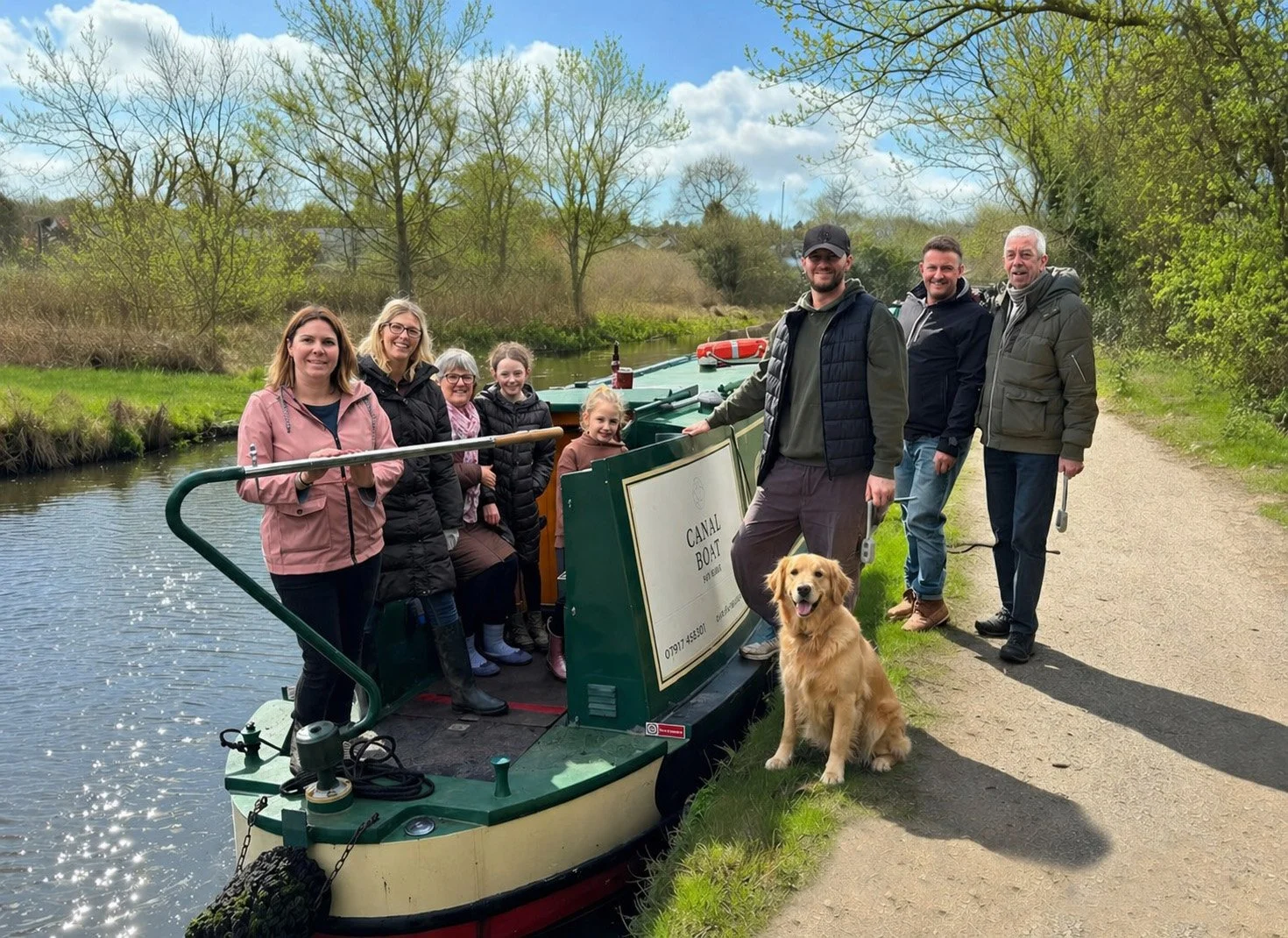 Group of people standing on a canal boat and beside a canal in Apsley, smiling outdoors in a green, wooded area on a cloudy day. A dog sits in front of them.