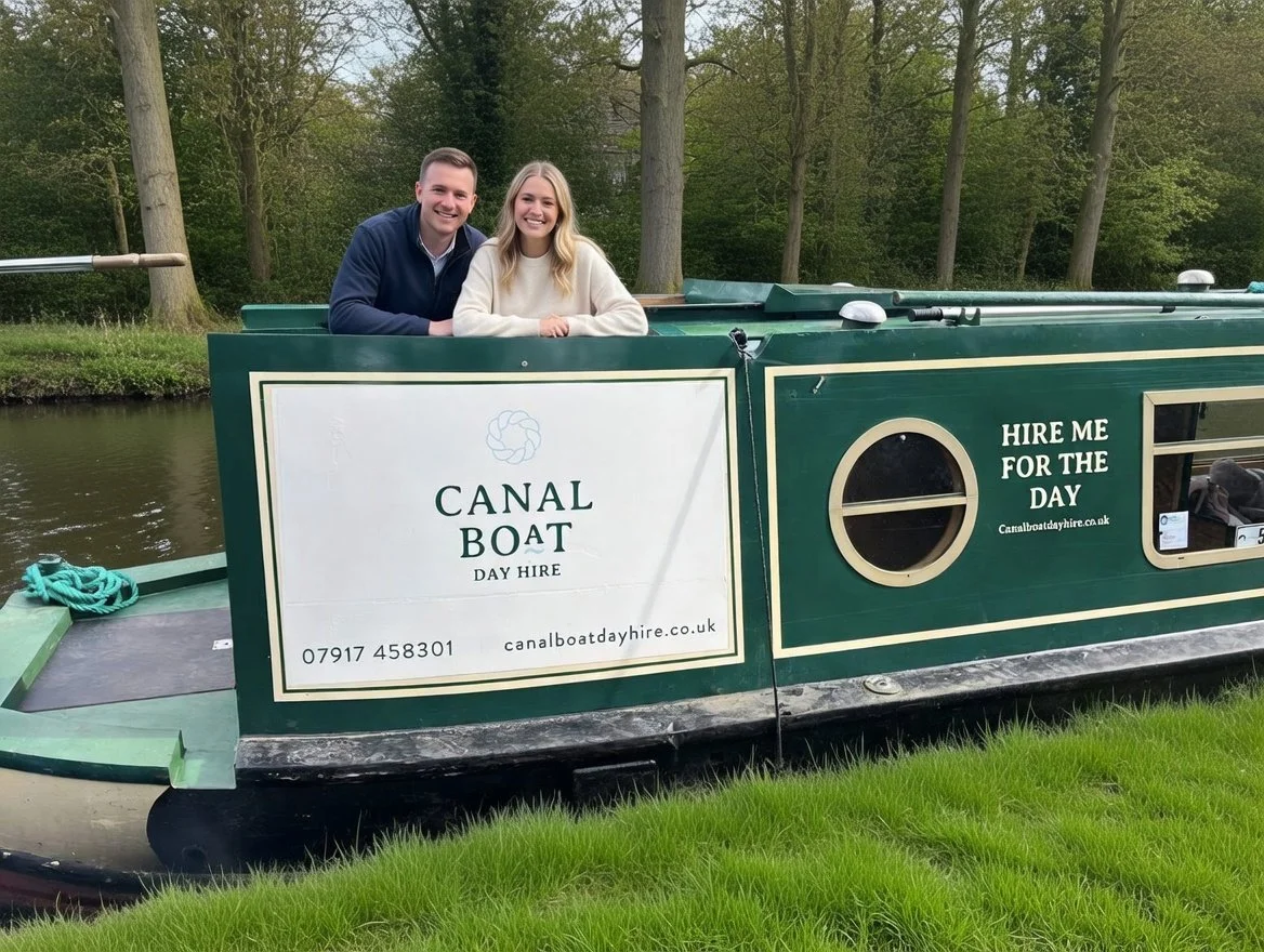 Two smiling people, a man and a woman, sitting on Tilly the canal boat on the Grand Union Canal surrounded by trees.