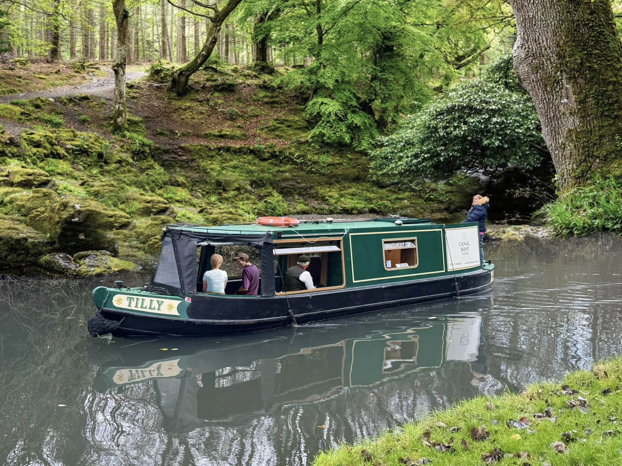 A narrowboat named Tilly is navigating a calm, narrow river in a lush green forest. The boat has a green and black exterior with a family of three and a captain on board, and one person standing at the stern. The riverbank is covered with grass and fallen leaves, with moss-covered rocks and tall trees surrounding the scene.