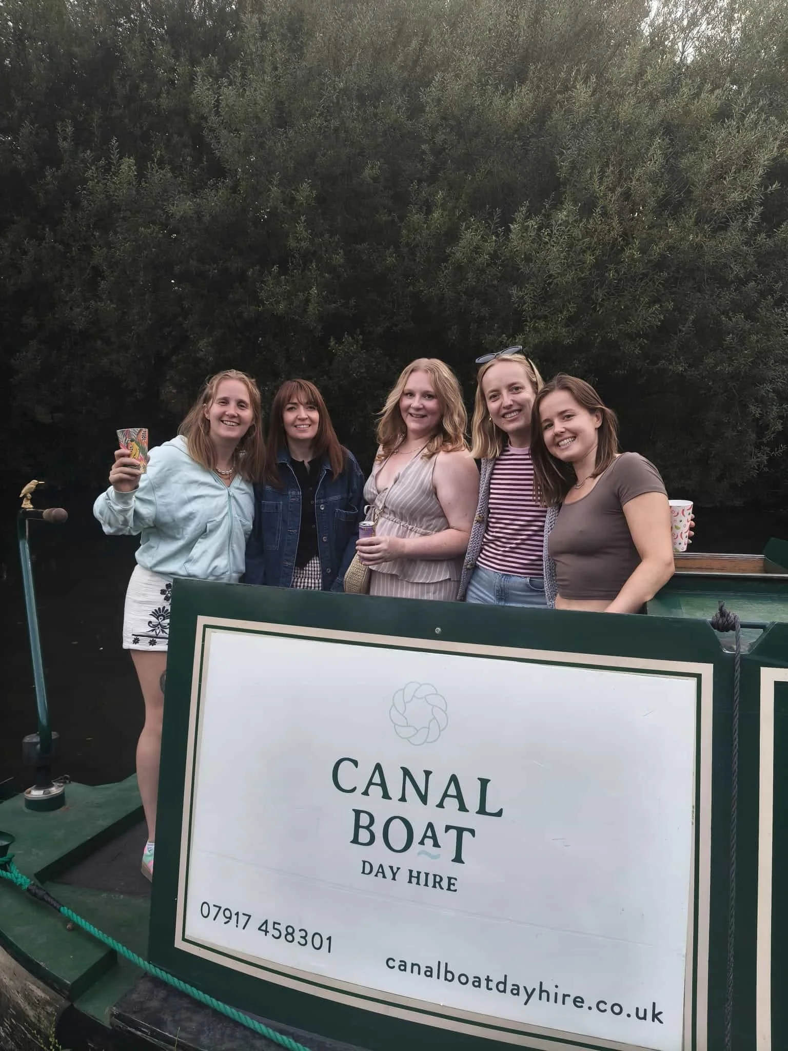 A group of five women standing together outdoors, smiling at the camera. They are gathered behind a sign that reads 'Canal Boat Day Hire' with contact information. They are in front of leafy greenery.
