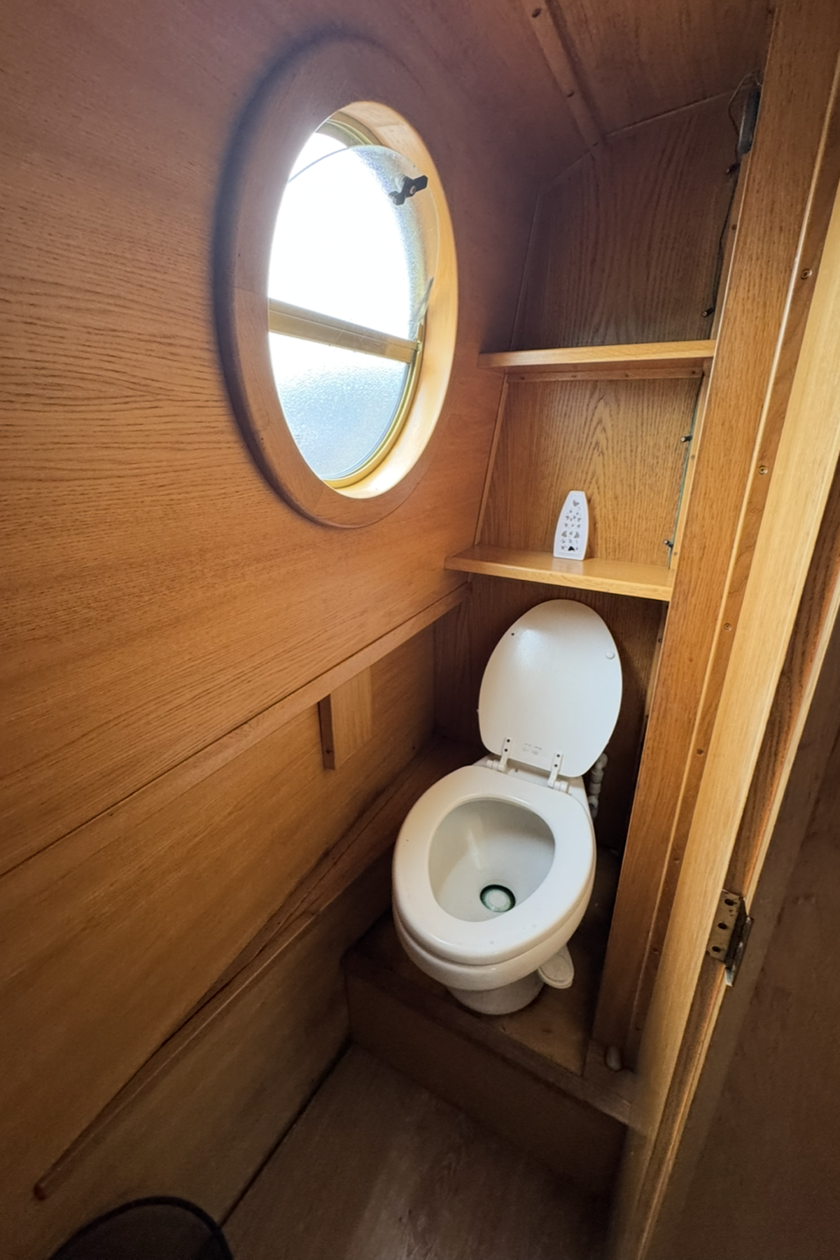 Wood-paneled bathroom with a toilet beneath an oval window, a small shelf, and a white air purifier on the shelf.