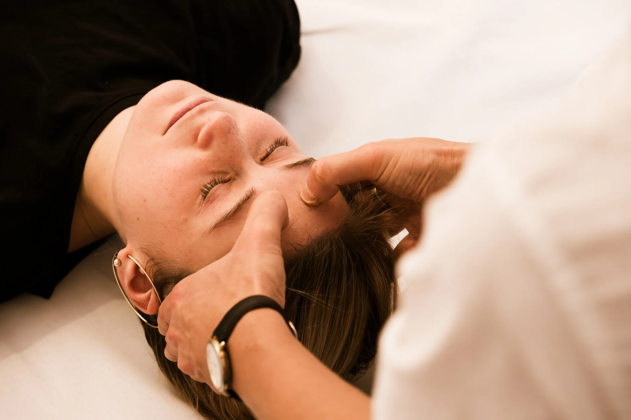 Person lying on their back receiving a Shiatsu massage from a therapist, who is using their fingers to massage the person's face. The person has closed eyes and is relaxed.