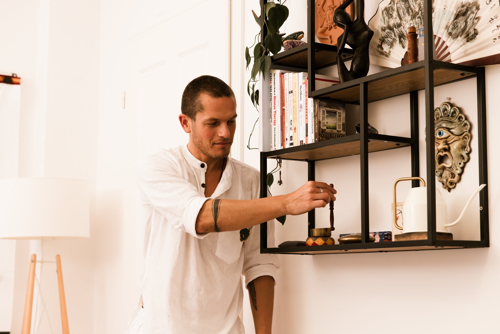 A man with short dark hair and a white shirt arranging objects on a black metal and wood shelving unit, decorated with various items including books, sculptures, a mask, and a potted plant, in a well-lit room with white walls.