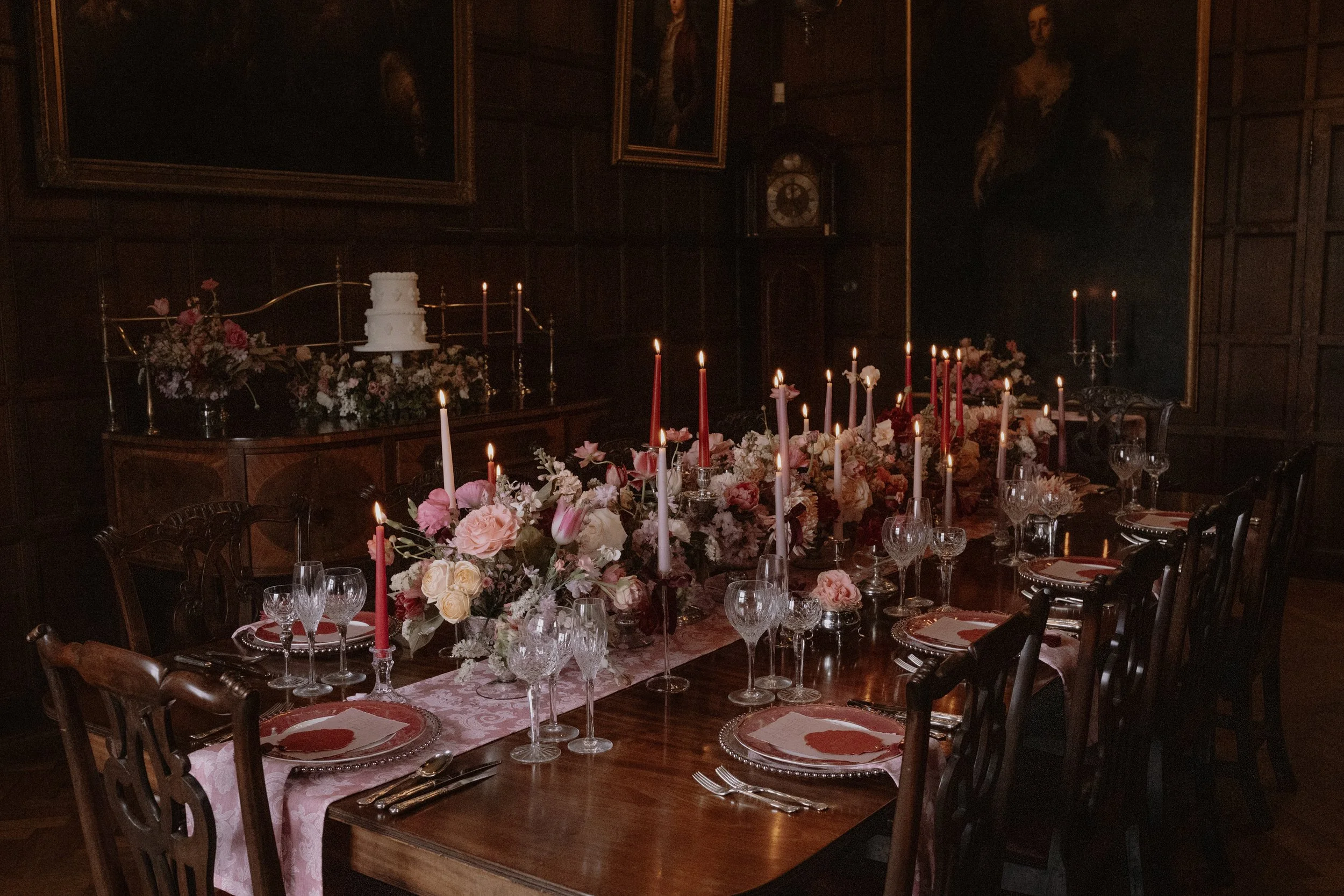 An antique dining room table is covere with a pink table running and place setting with a centre of flowers in pale and dark pinks with lots of candlesticks