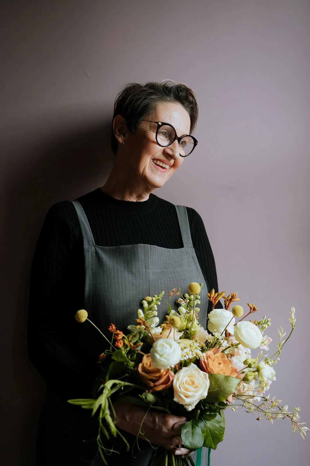 A smiling woman with short gray hair and glasses holding a wild, natural bouquet of various flowers, standing against a plain light purple wall.