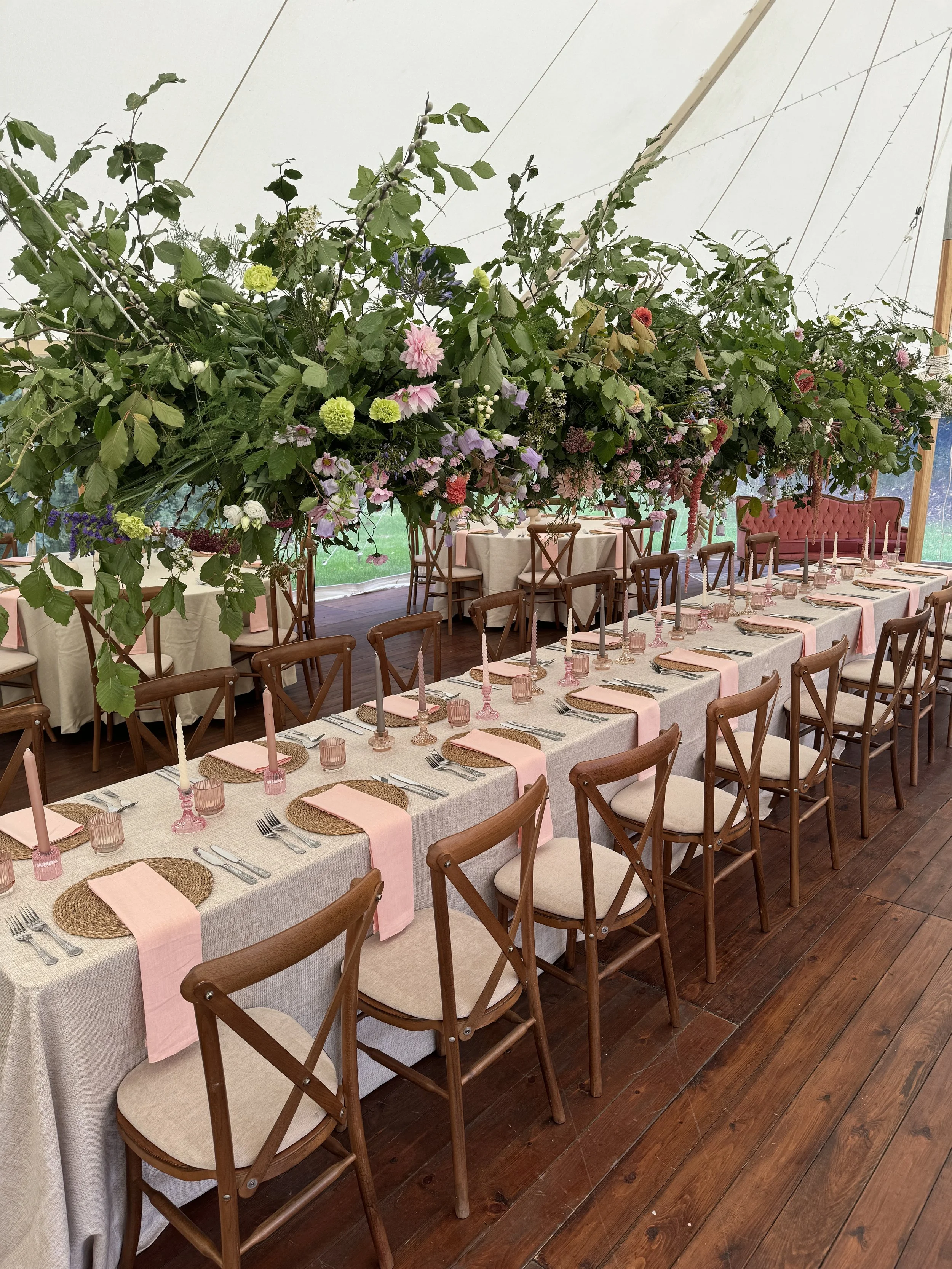 Long dining table decorated with pink napkins, candles, and pink glasses, set for a formal event with wooden chairs around and covered with a beige tablecloth, topped with a large floral arrangement.