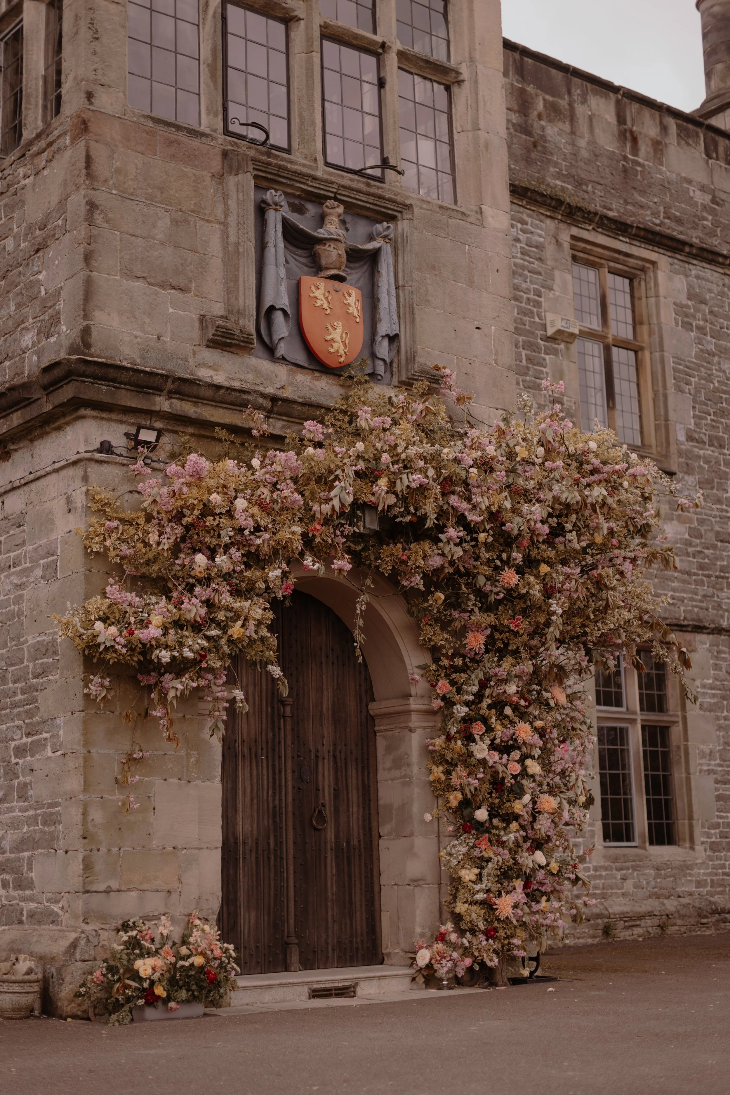 A historic stone building with an arched wooden door covered in pink and white flowers, a coat of arms crest above the door, and stone windows.