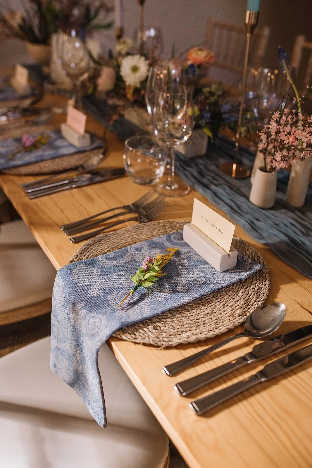 A decorated dining table set for a formal event with place settings, floral centerpieces, and a blue patterned napkin with a sprig of greenery.