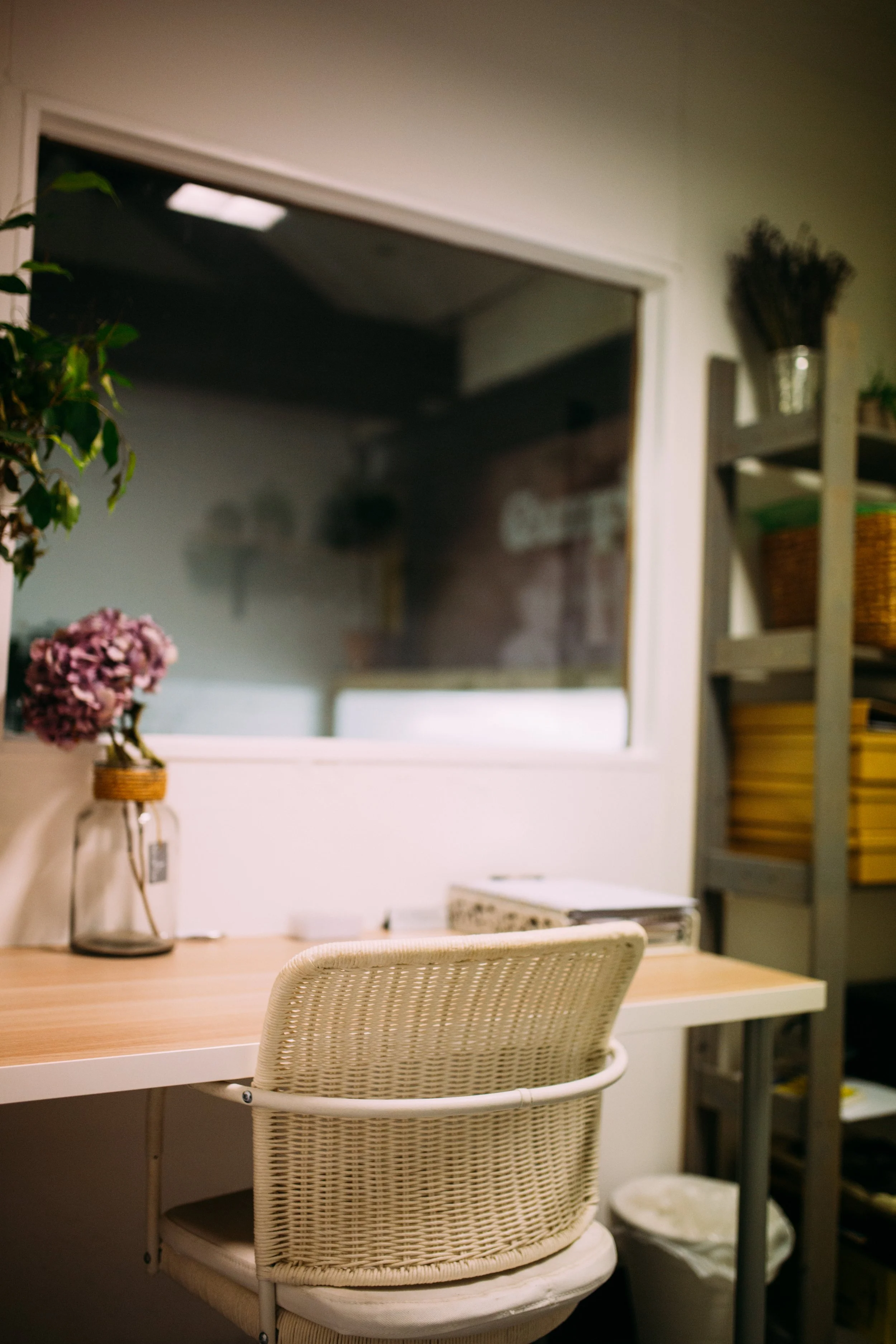 A cozy office desk with a wicker chair, a vase with pink flowers, and shelves with baskets and yellow trays.