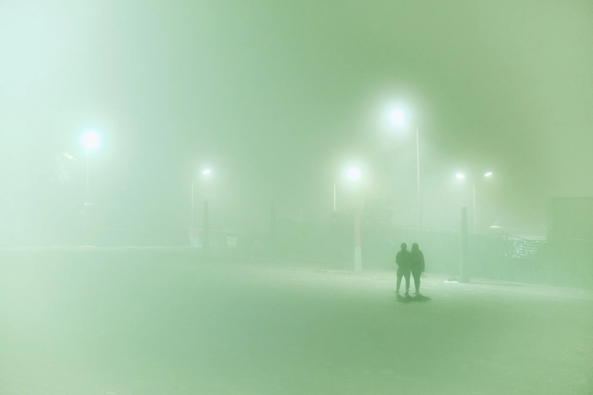 Dos personas caminando en una zona con niebla o humo, con farolas iluminando y un ambiente oscuro.