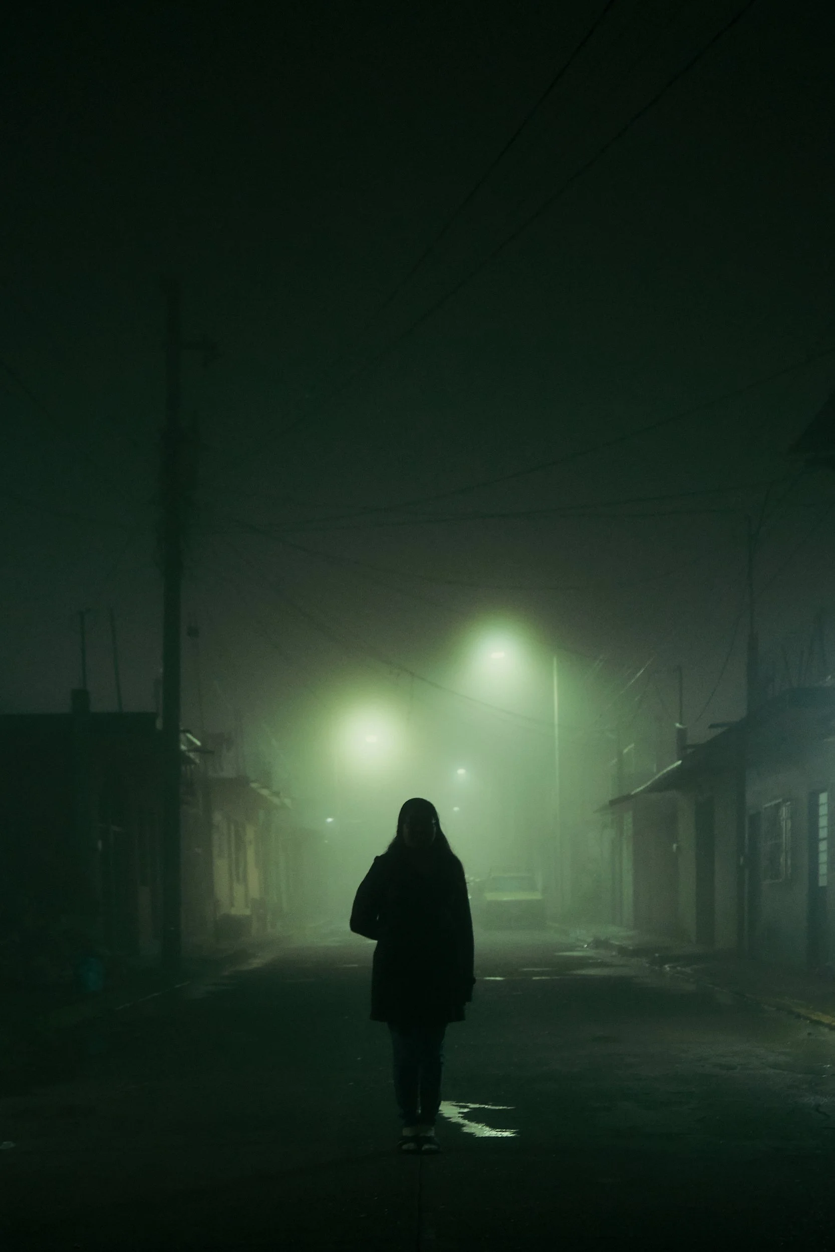 Una persona caminando en una calle nocturna con neblina, luz verde y postes de luz, en un entorno oscuro y misterioso.