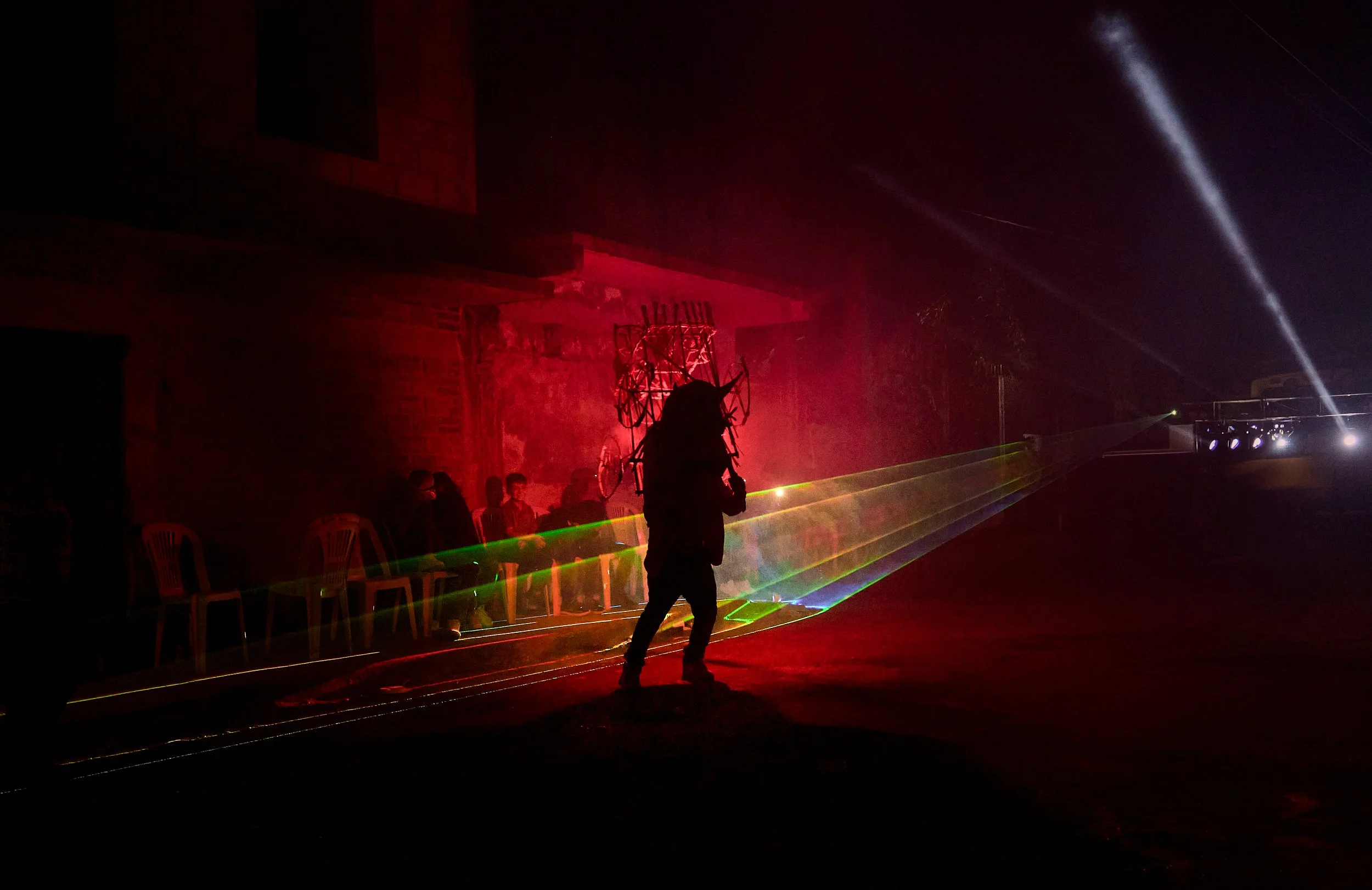 Personas paseando en un evento nocturno con luces de láser y decoraciones luminosas, en un entorno al aire libre con sillas de plástico y madera.