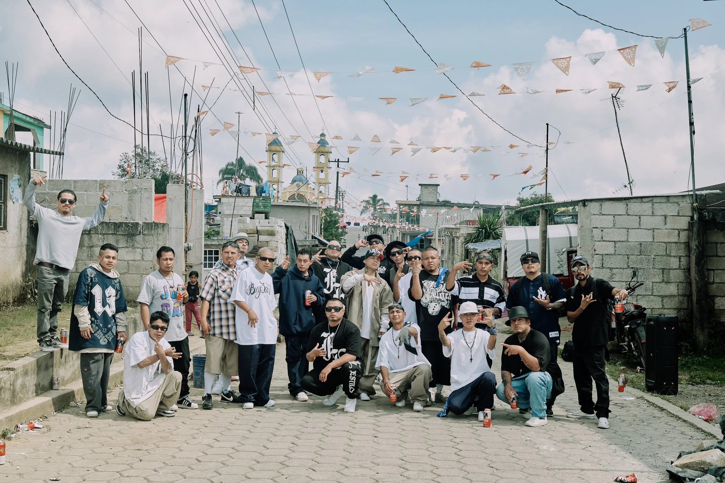 Grupo de personas posando en una calle de barrio, algunas con camisetas y gorras, con banderines decorativos en el cielo y construcciones de ladrillo, mientras algunos sostienen bebidas en lata.
