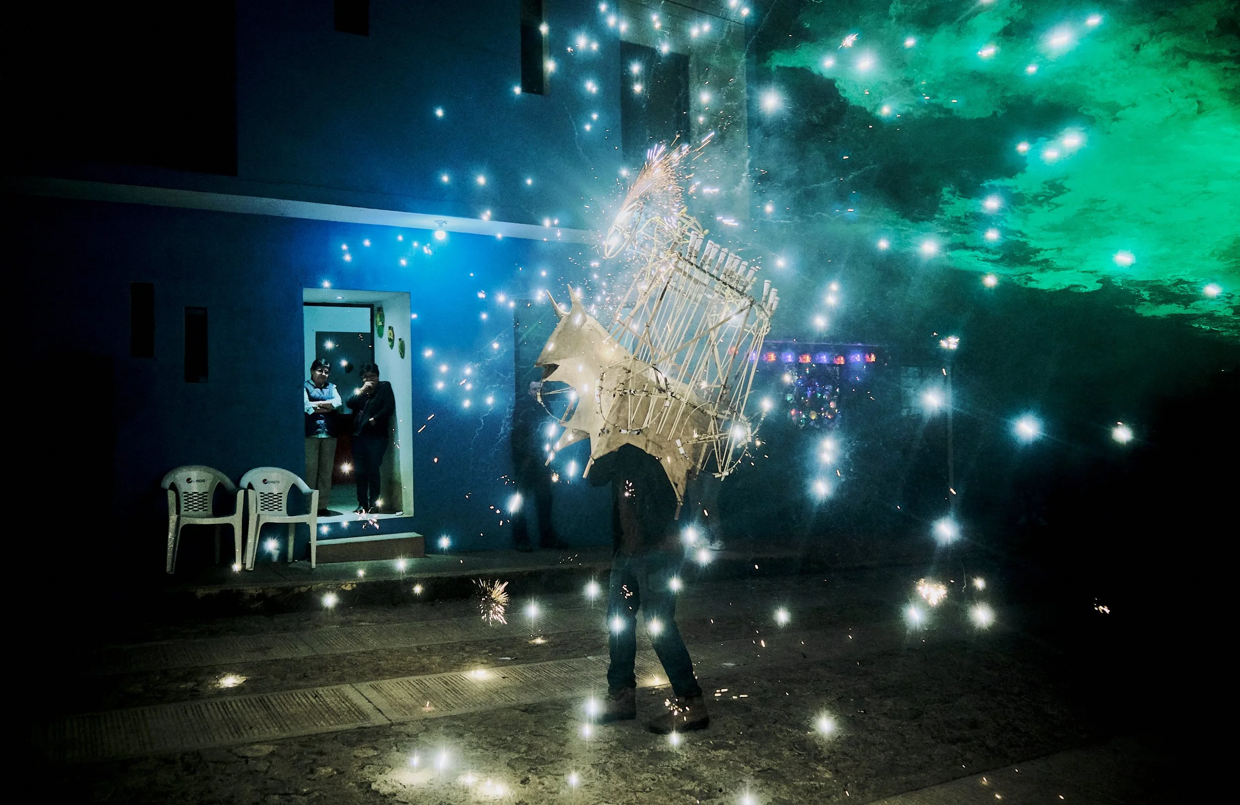 Celebración con fuegos artificiales y shows de luces en una fiesta o evento nocturno, con varias personas observando el espectáculo cerca de una estructura decorativa.