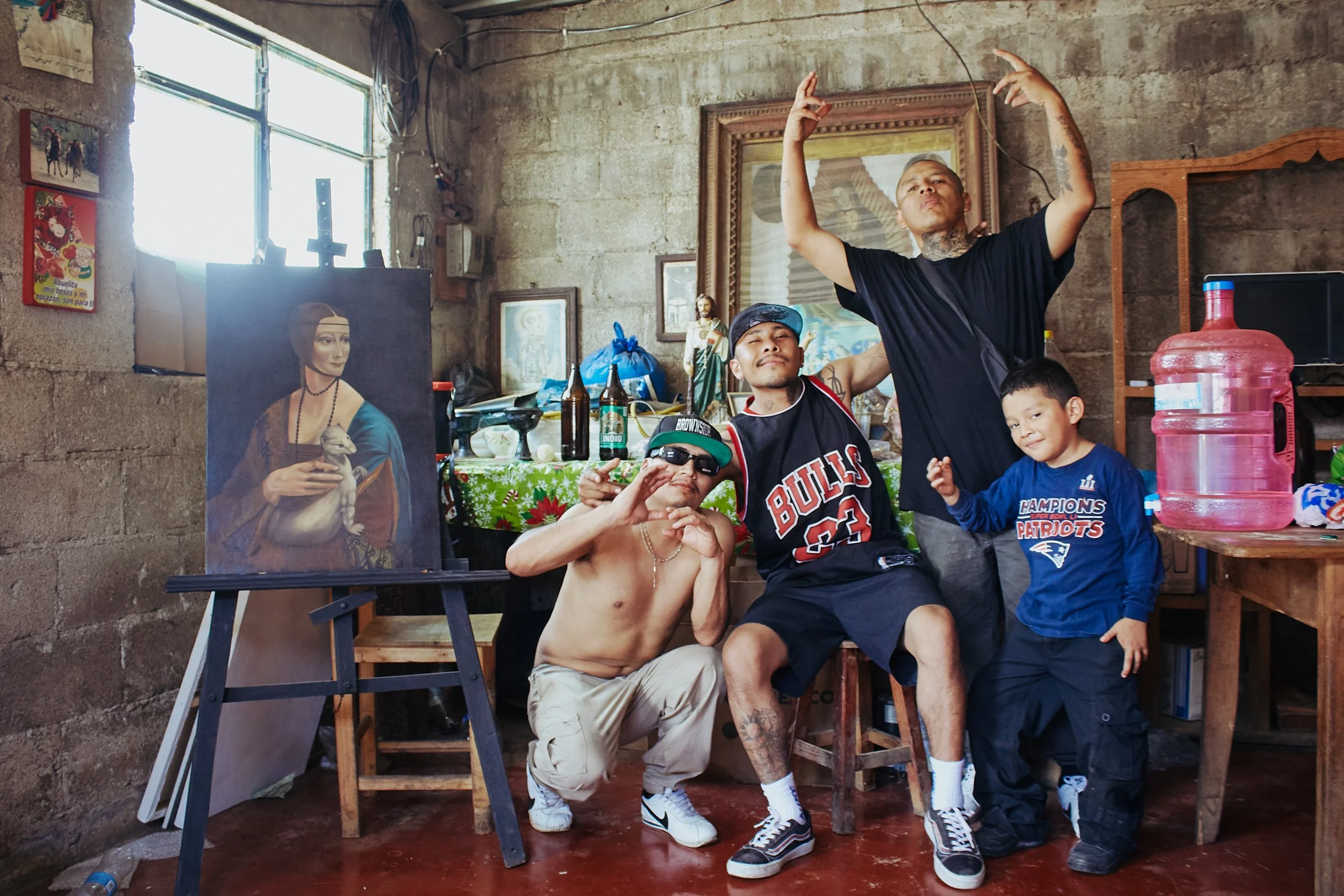 Grupo de personas en una habitación con paredes de ladrillo, algunos niños y un joven con tatuajes y cabello rapado posando junto a una pintura de una mujer con sombrero, en un entorno informal y rústico.