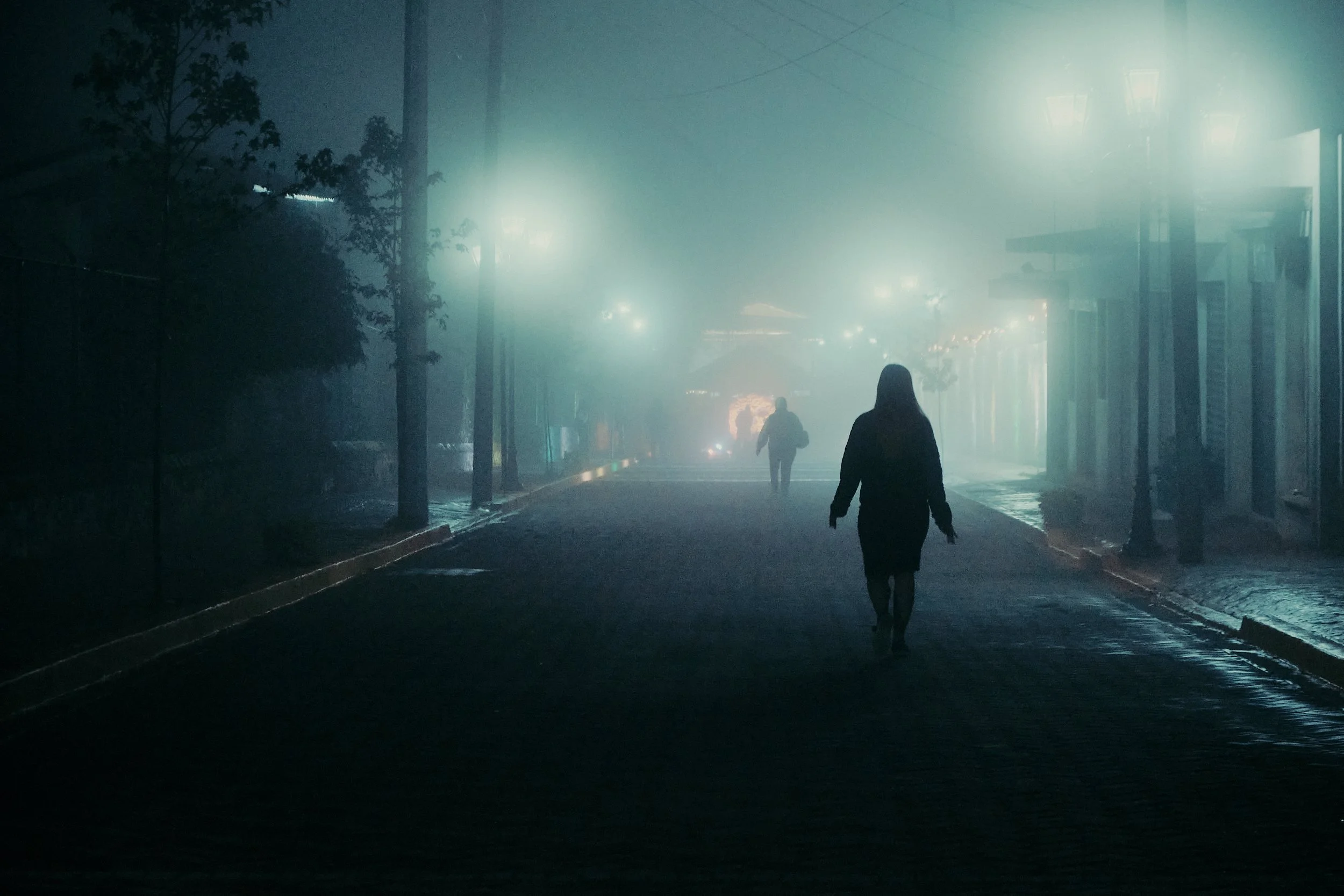 Paseo nocturno por una calle nebulosa iluminada por faroles, con dos personas caminando.