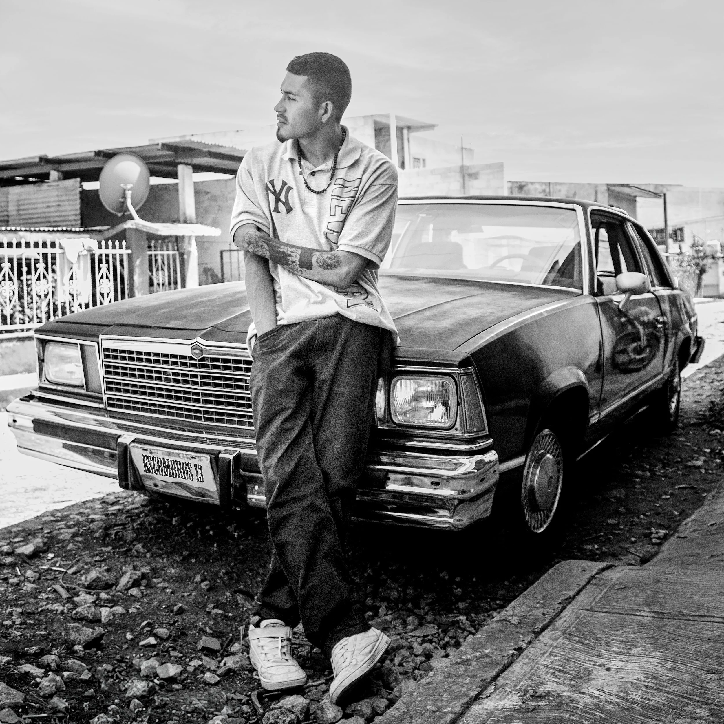 Joven con tatuajes y cadenas, con ropa deportiva, apoyado en un coche clásico, en un ambiente urbano con edificios y un satélite en el fondo, en blanco y negro.