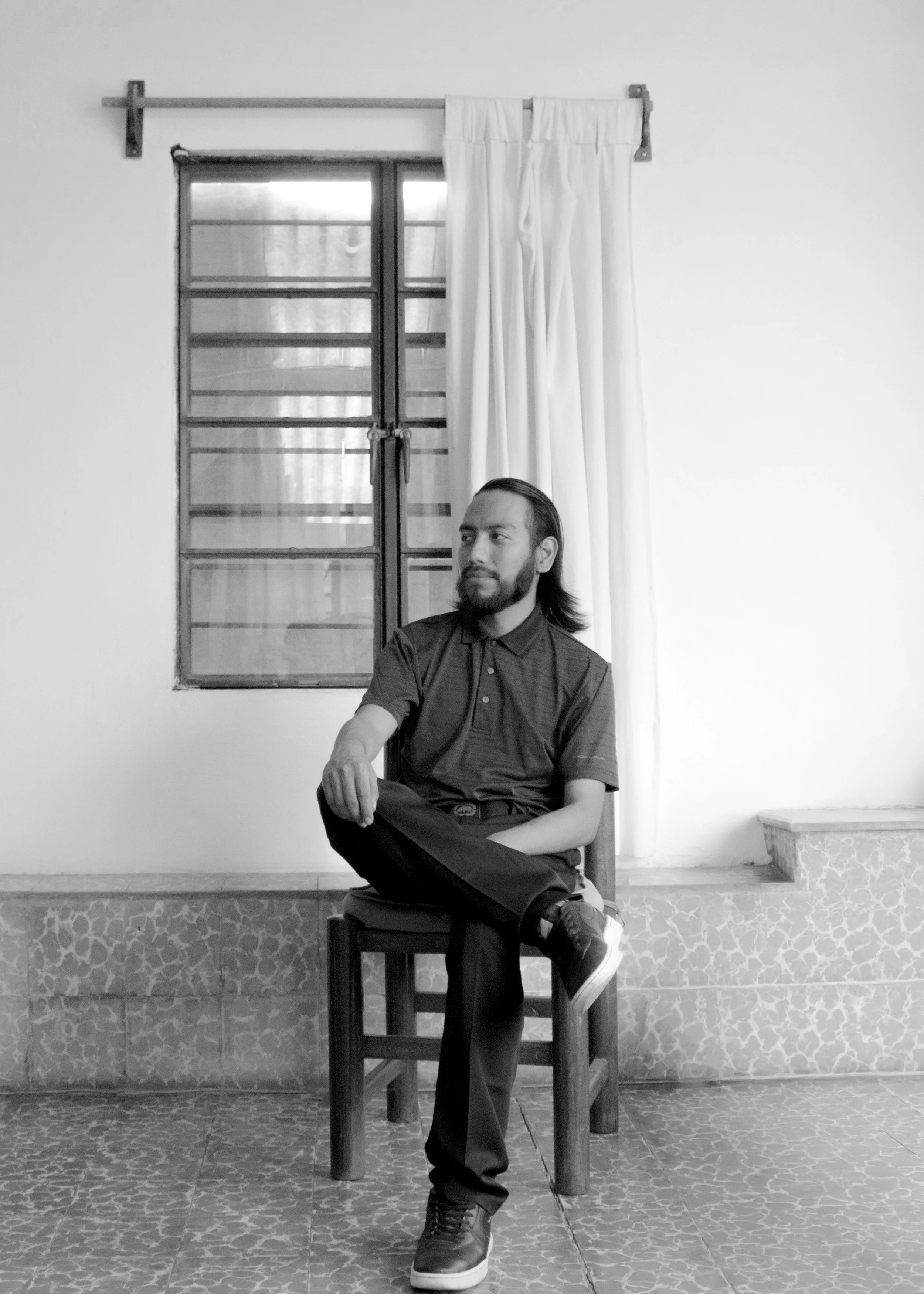Un hombre con barba y cabello largo sentado en una silla de madera, llevando puesta una camisa gris y pantalones oscuros, en un entorno interior con pared blanca, cortinas blancas y una ventana con rejas metálicas.