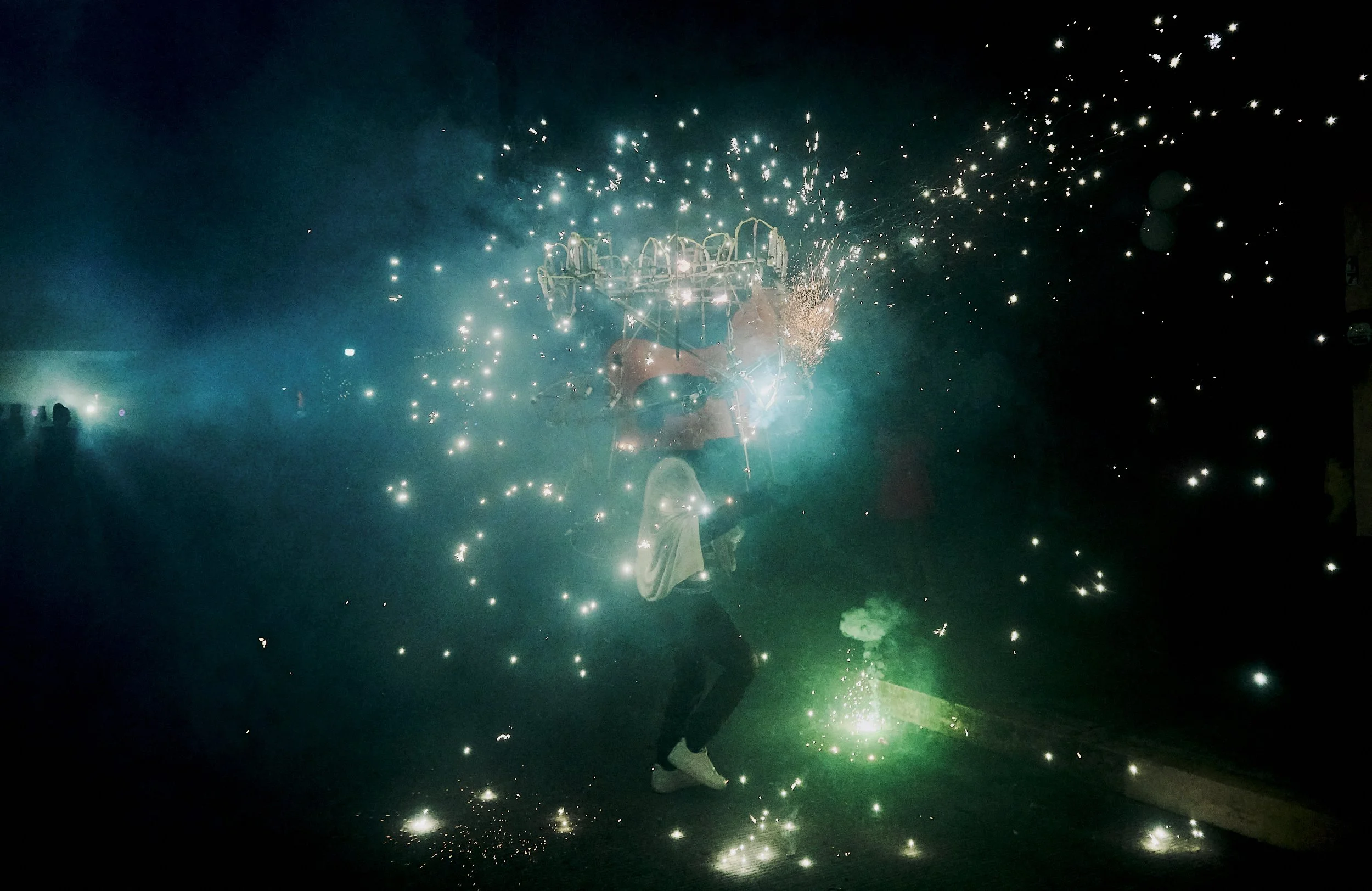 Una persona con ropa clara sosteniendo un objeto que emite chispas y humo, en medio de fuegos artificiales que iluminan el cielo en la noche.