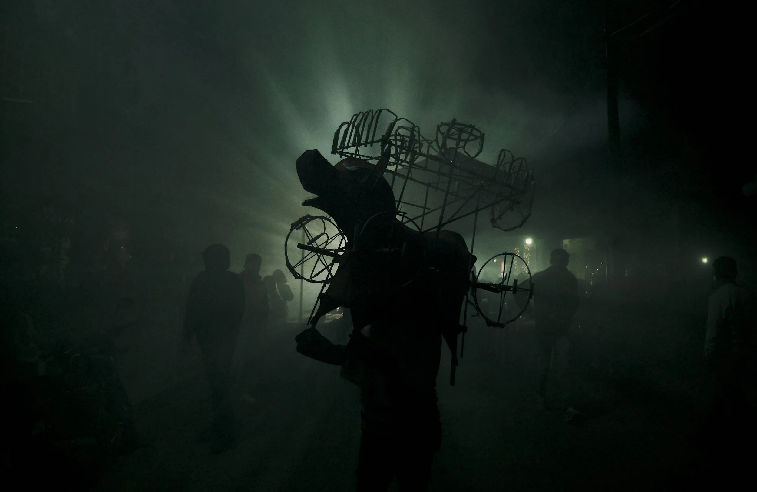 Persona sosteniendo una estructura metálica en un ambiente oscuro con humo y siluetas de personas al fondo.