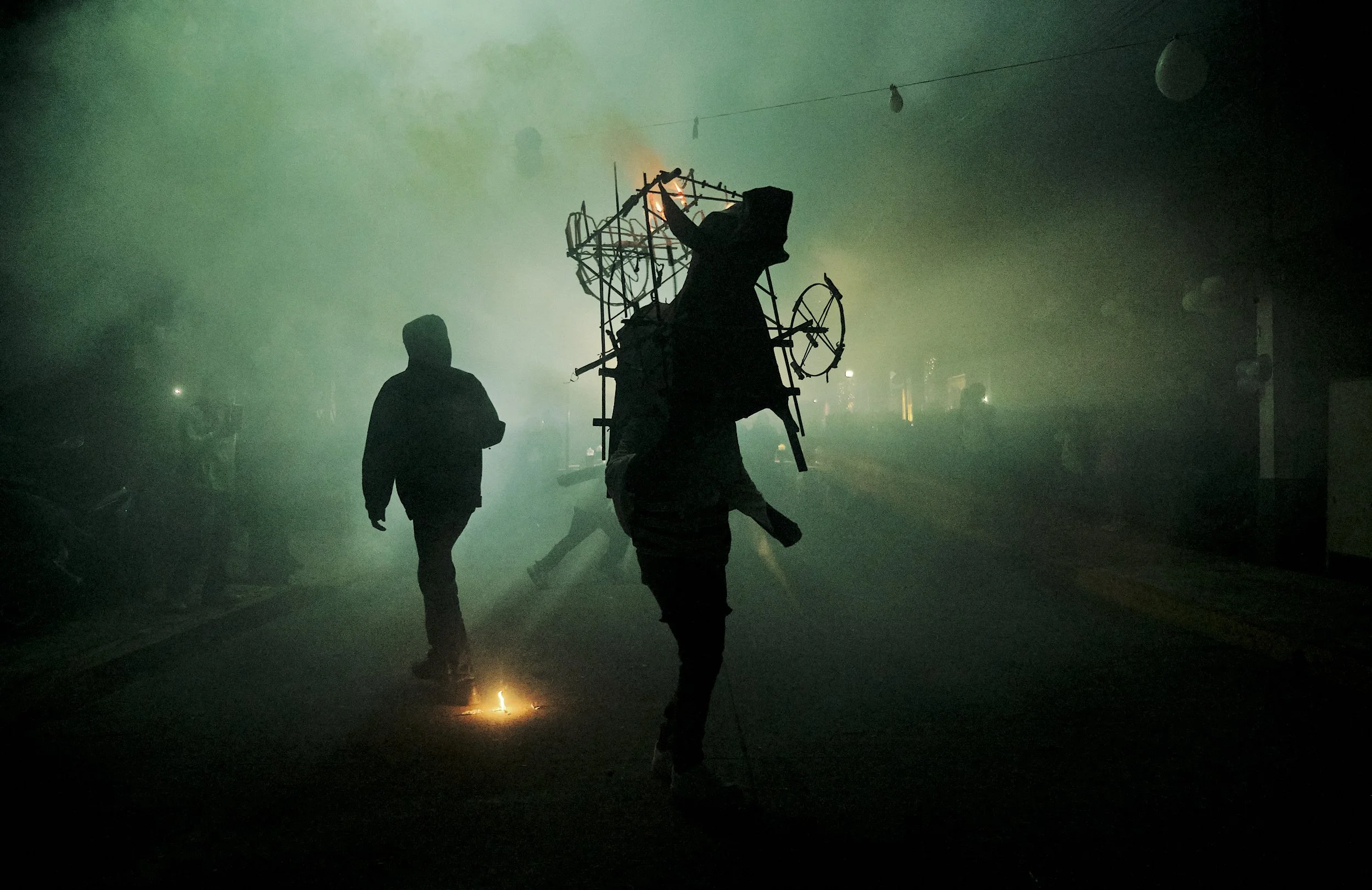 Persona con capucha que camina en una calle obscura, con humo y luces, y otra persona llevando una estructura metálica