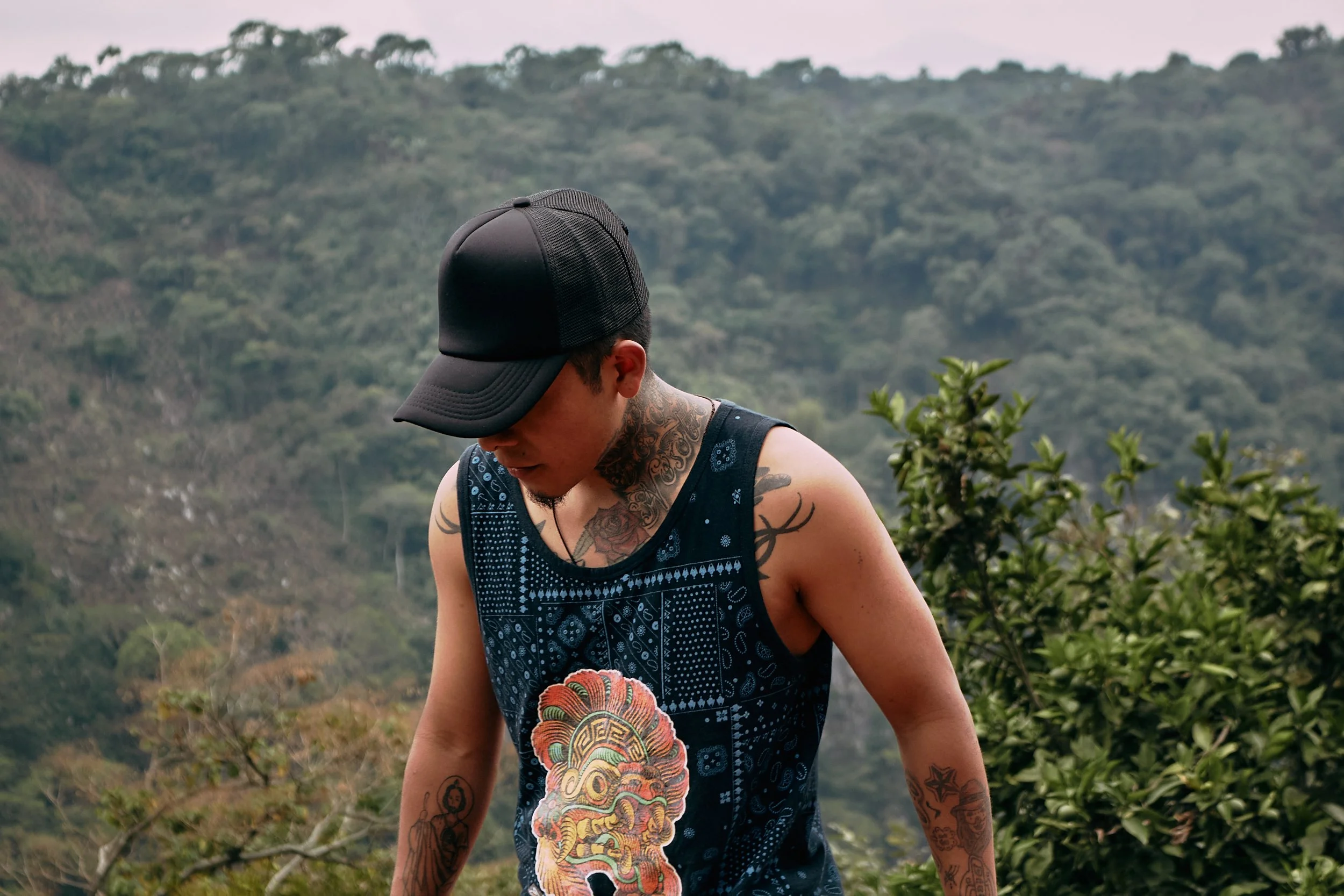 Joven con tatuajes y gorra negra, mirando hacia abajo, en un entorno natural con vegetación y colinas.