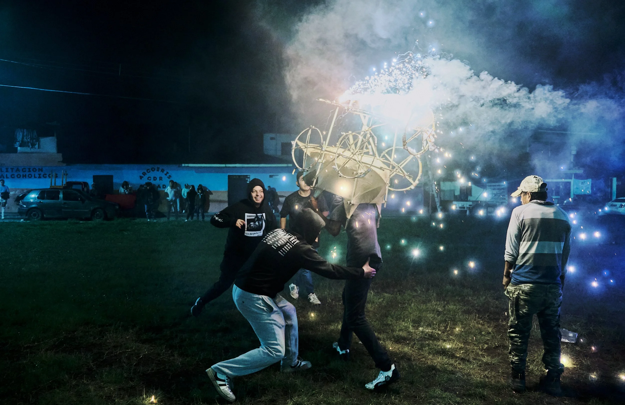 Personas celebrando con fuegos artificiales en un espacio abierto de noche, algunos sosteniendo cómo un chiflado de fuego y otros observando.