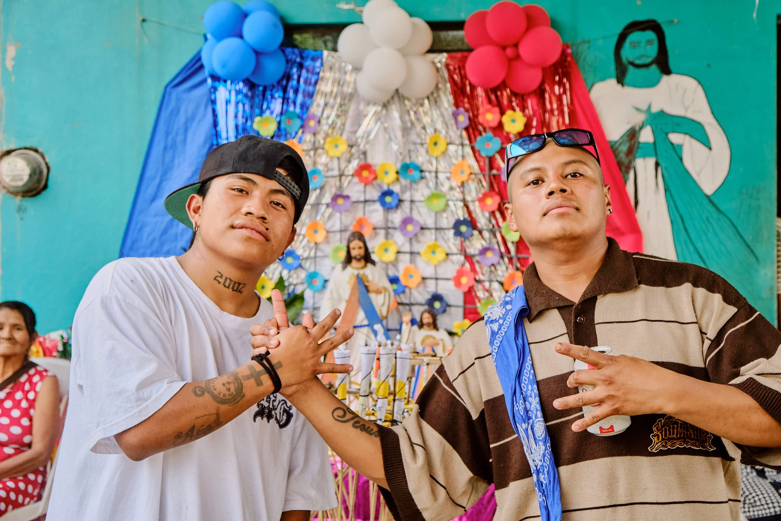 Dos jóvenes hombres posando en una celebración con decoración colorida, globos y figuras religiosas, ambos mostrando signos de paz con las manos.