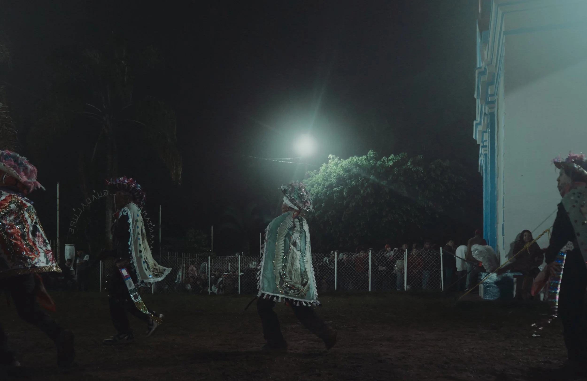 Personas participando en una danza tradicional al aire libre en la noche, usando trajes coloridos y sombreros decorativos, con un grupo de espectadores al fondo.