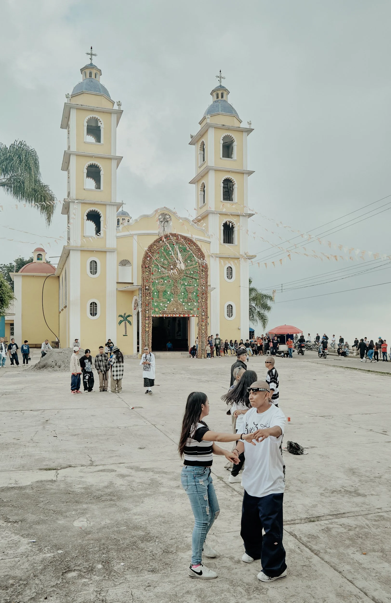 Una iglesia con dos torres y decoraciones coloridas frente a una plaza con muchas personas bailando y caminando, en un día nublado.