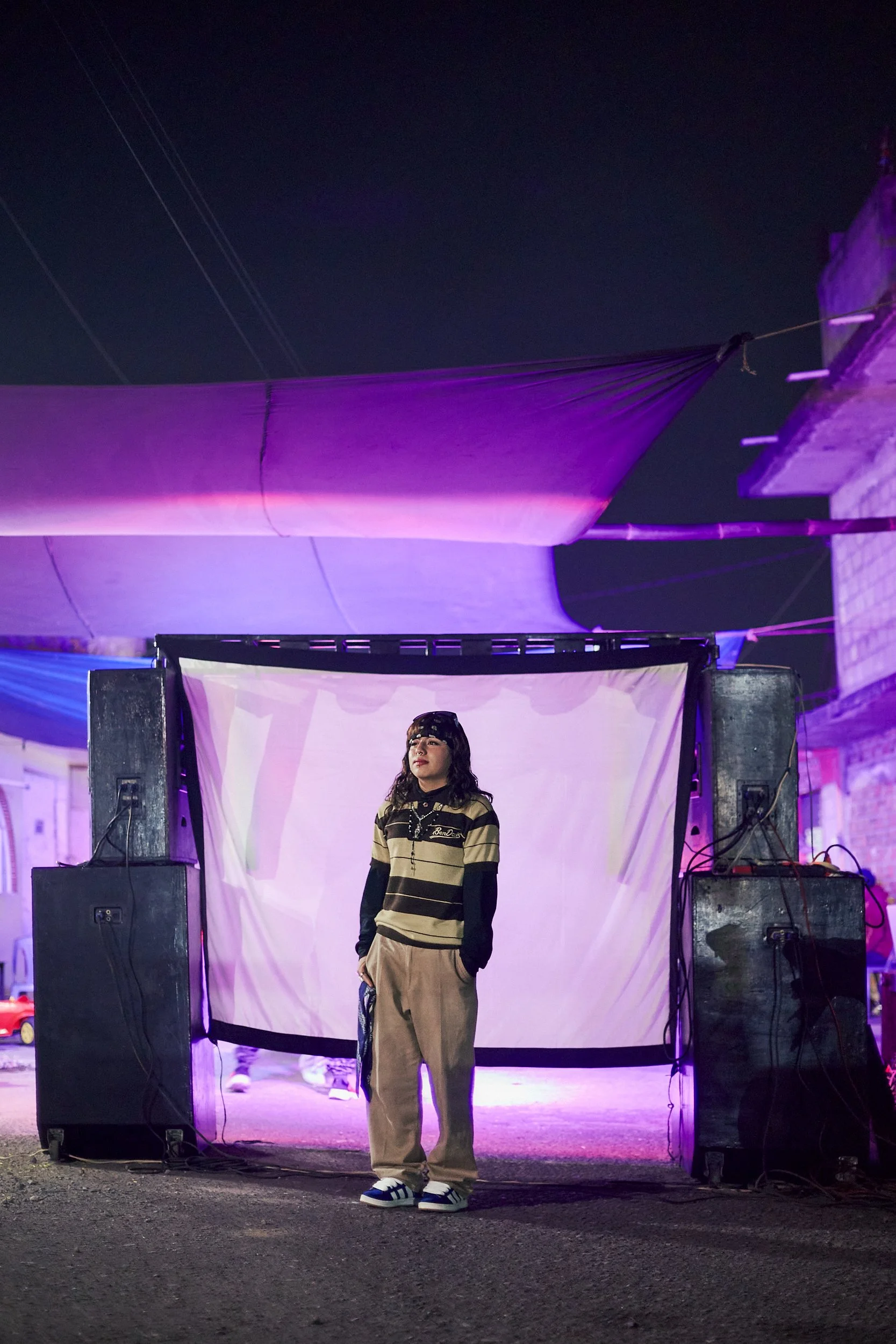 Joven con ropa casual, de pie frente a una pantalla en un escenario, con sonido y cables, en un evento nocturno con decoraciones con luces violetas.