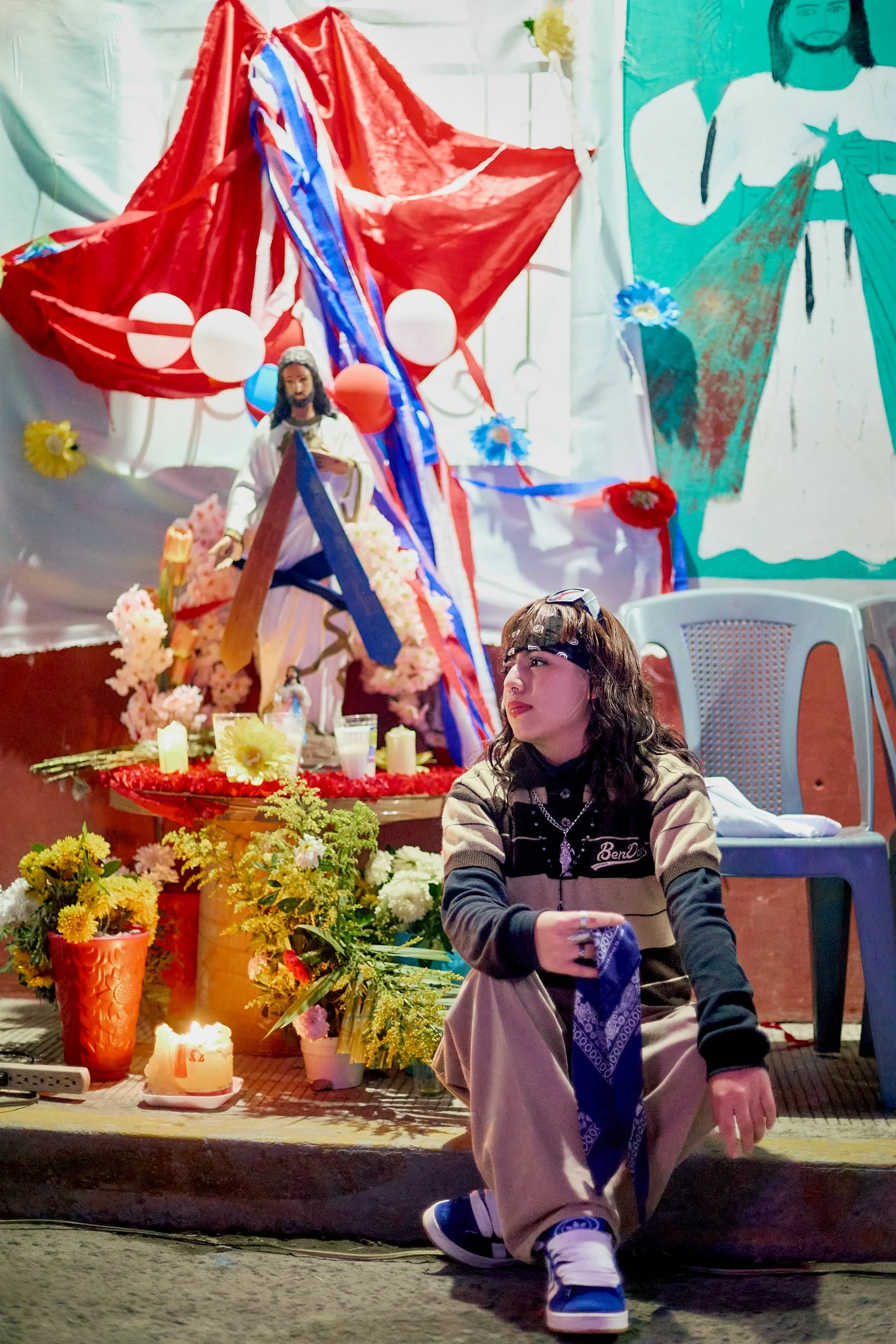Escena de celebración religiosa con ofrendas florales, velas, y una figura de Jesús en el altar, decorado con banderas y flores de colores, en un entorno que parece ser un altar casero o comunitario.