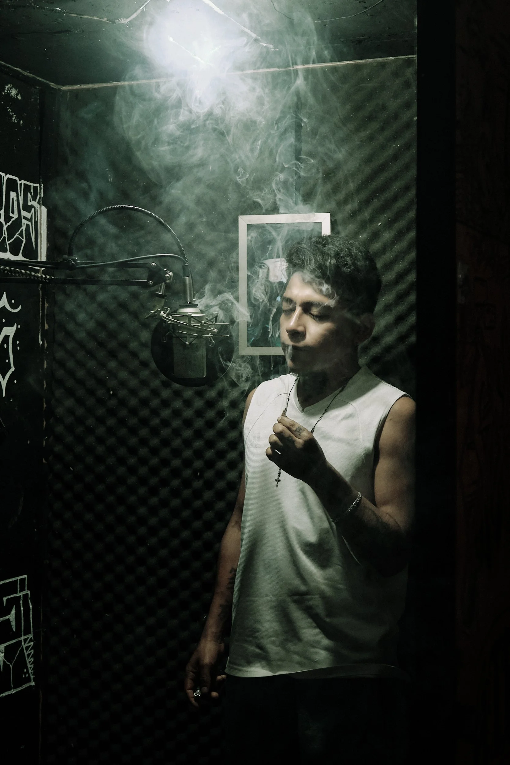 Joven en cabina de grabación, con auriculares y mascarilla, rodeado de paredes con paneles acústicos y humo, probablemente en una sesión de grabación musical o de voz.
