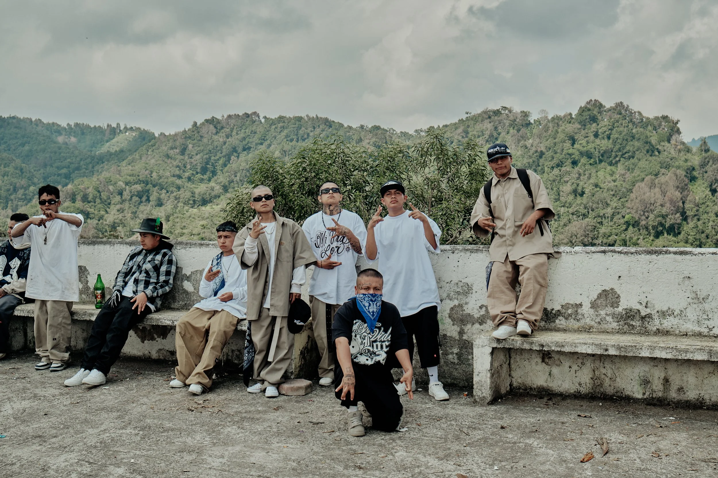 Grupo de jóvenes sentados y de pie en un muro, algunos con gafas de sol, en un entorno natural con montañas y cielo nublado al fondo.