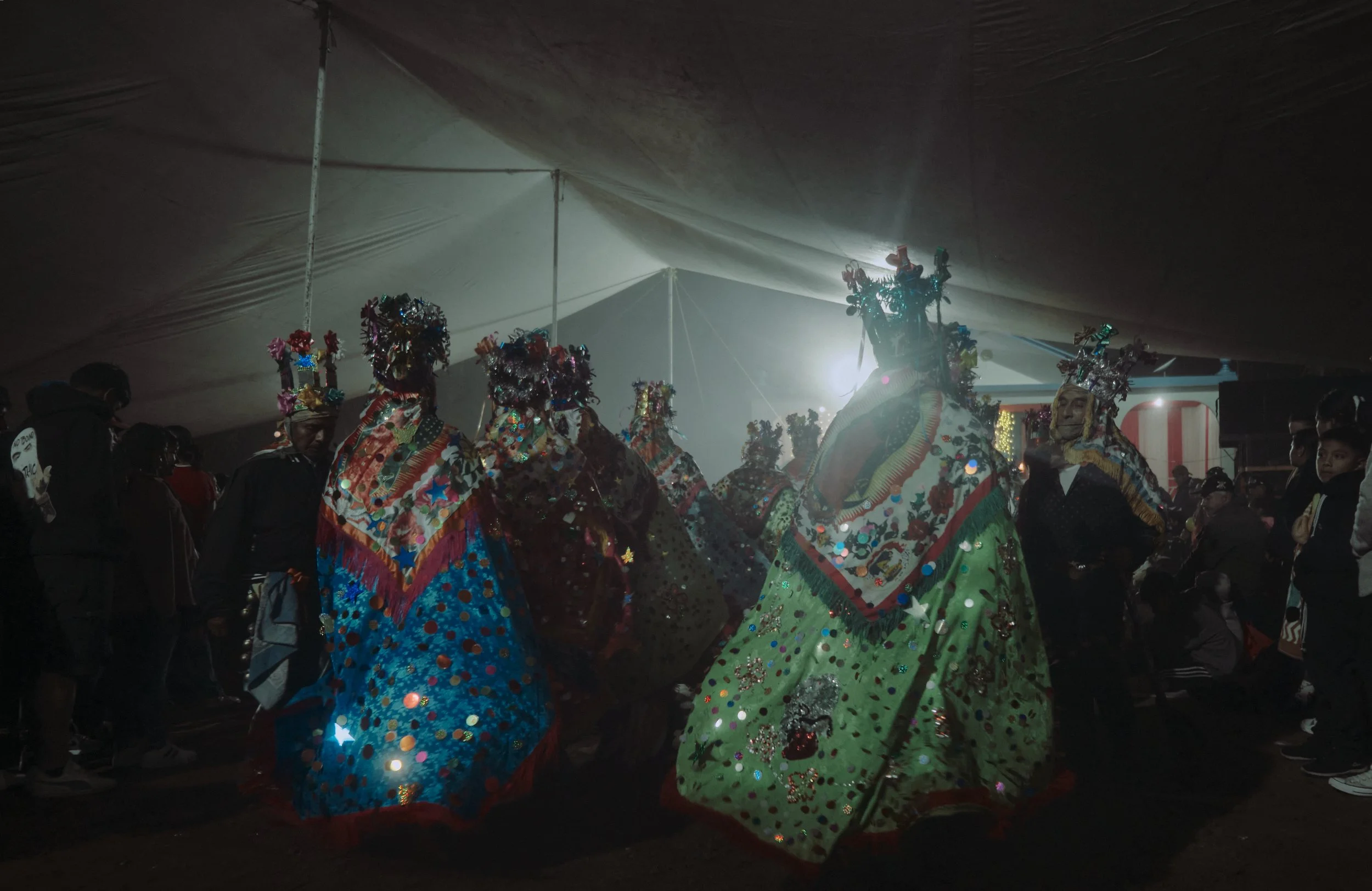 Personas bailando en una celebración tradicional, usando trajes coloridos con decoraciones y sombreros elaborados, en un ambiente con poca iluminación y luz brillante al fondo.