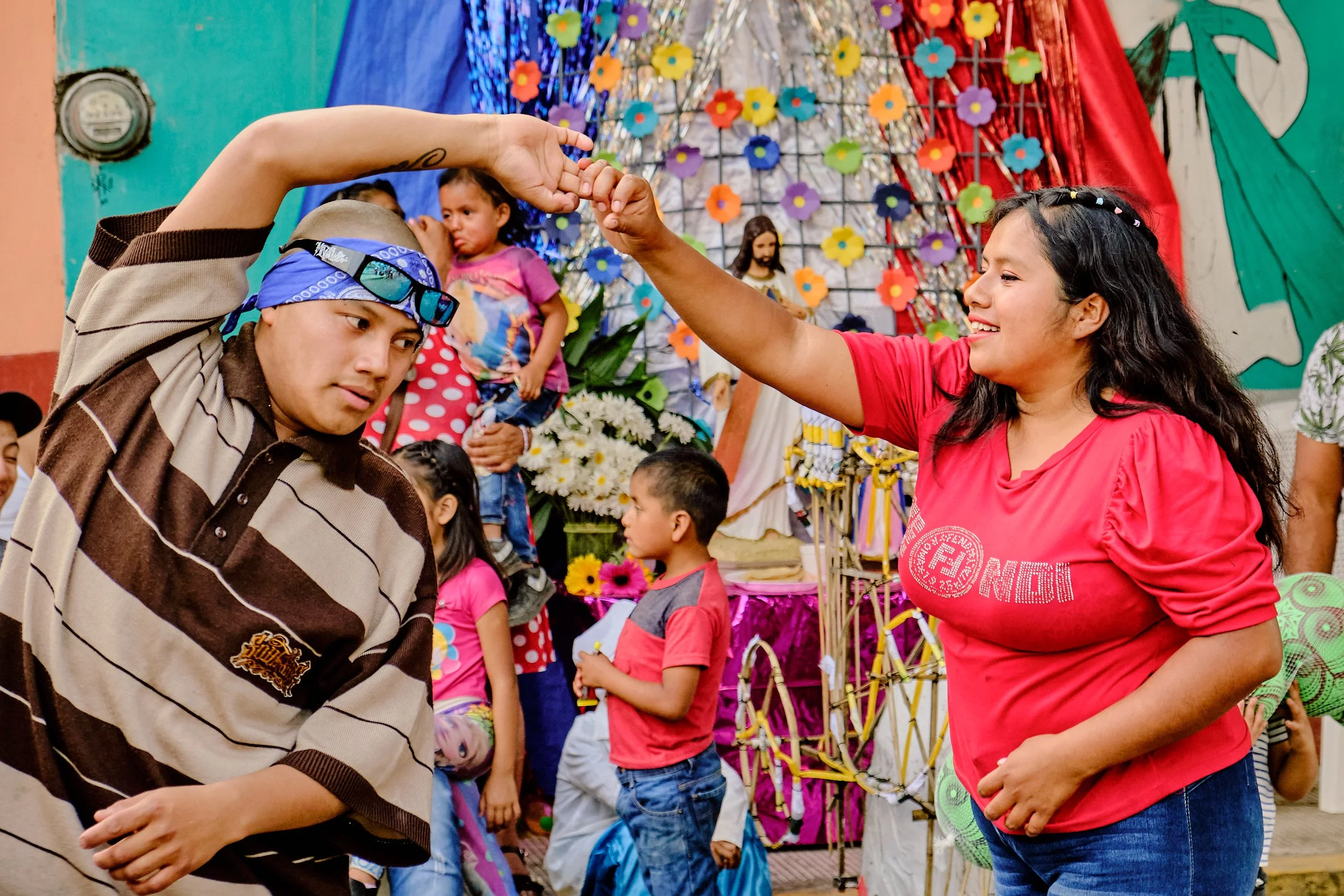 Personas participando en una celebración tradicional con decoraciones coloridas, una mujer y un joven bailando y niños en el fondo, en un ambiente festivo.