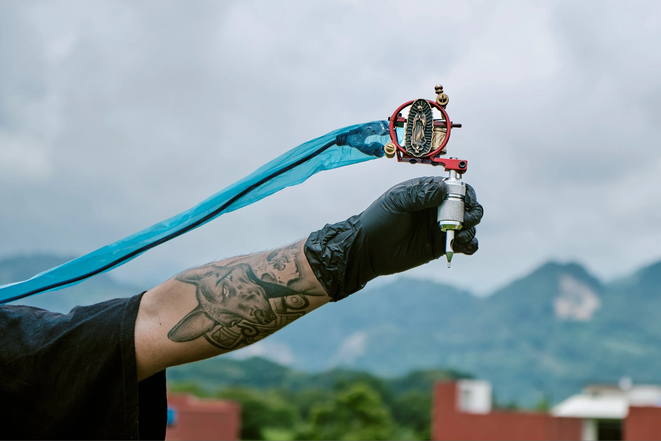 Una mano con tatuajes sosteniendo un selector de vinil, con un fondo de cielo nublado y áreas verdes y edificios en la distancia.
