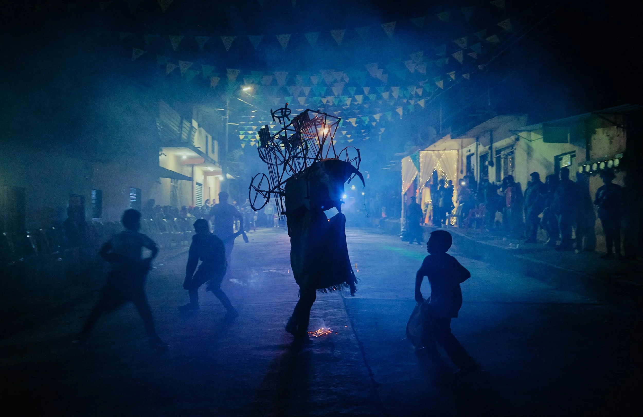 Una escena nocturna en una calle de un pueblo con muchas personas. En el centro, hay una persona con un sombrero que sostiene una estructura hecha de fierros y otros materiales, probablemente en una fiesta o celebración. La calle está decorada con ba