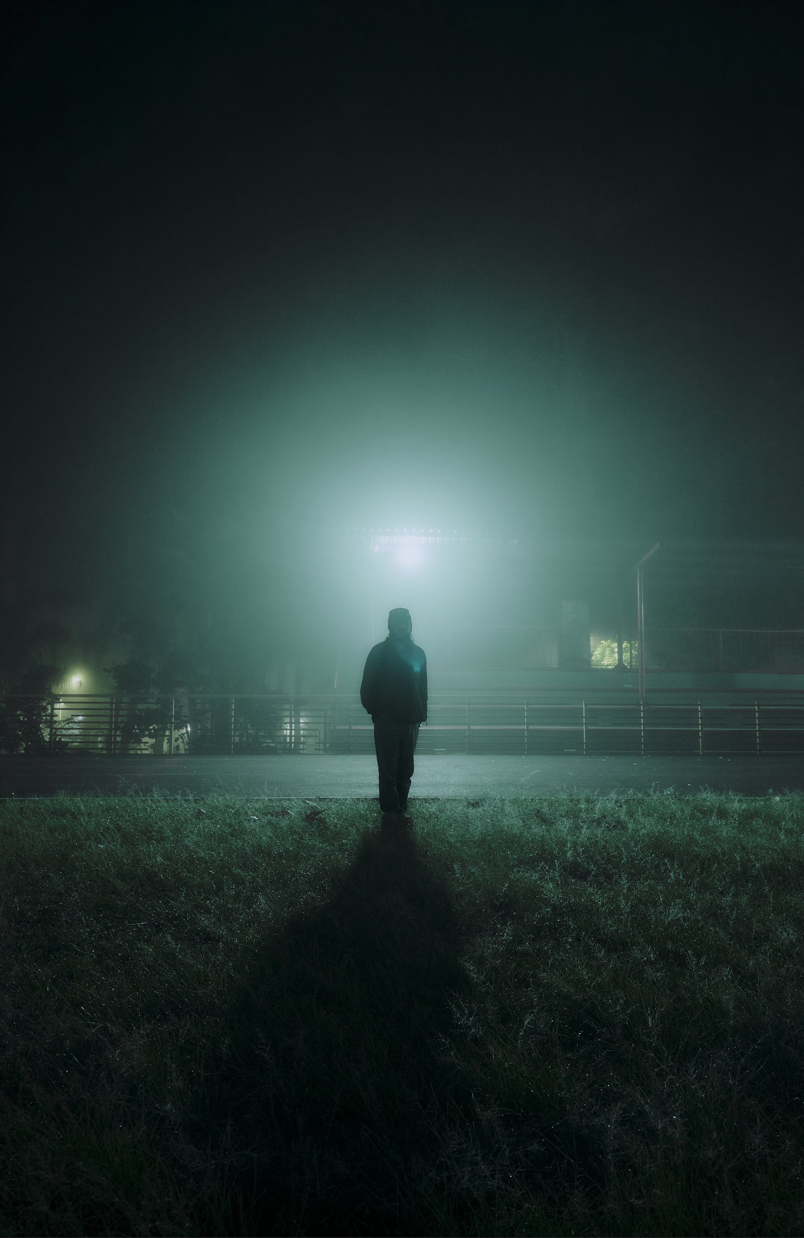 Persona caminando en un parque oscuro con niebla, iluminación intensa desde atrás, árboles y cercas visibles.