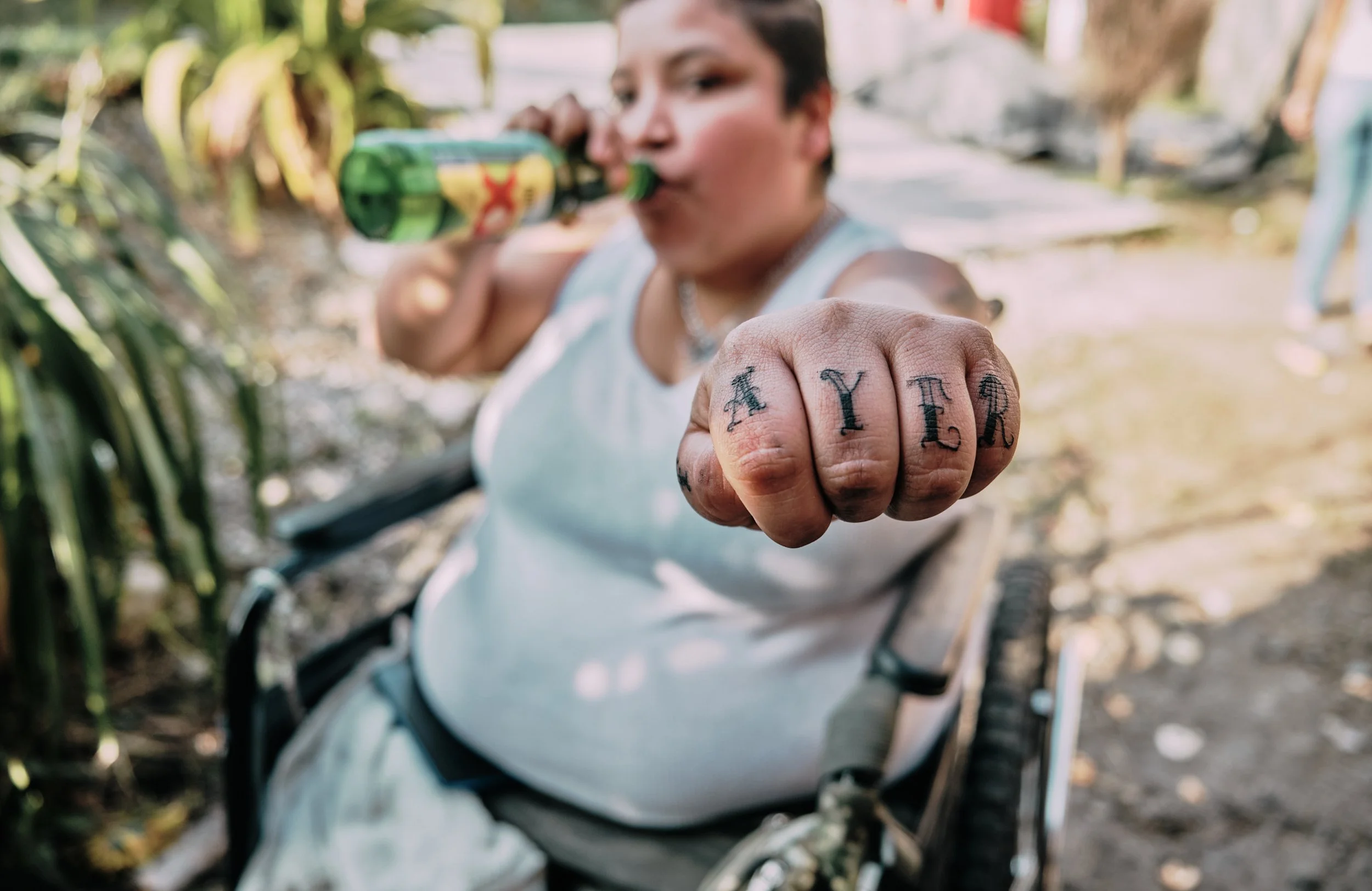 Persona con tatuajes en los dedos mostrando la palabra 'ALER' con un dedo, mientras sostiene una botella con una mano en una zona verde y soleada.