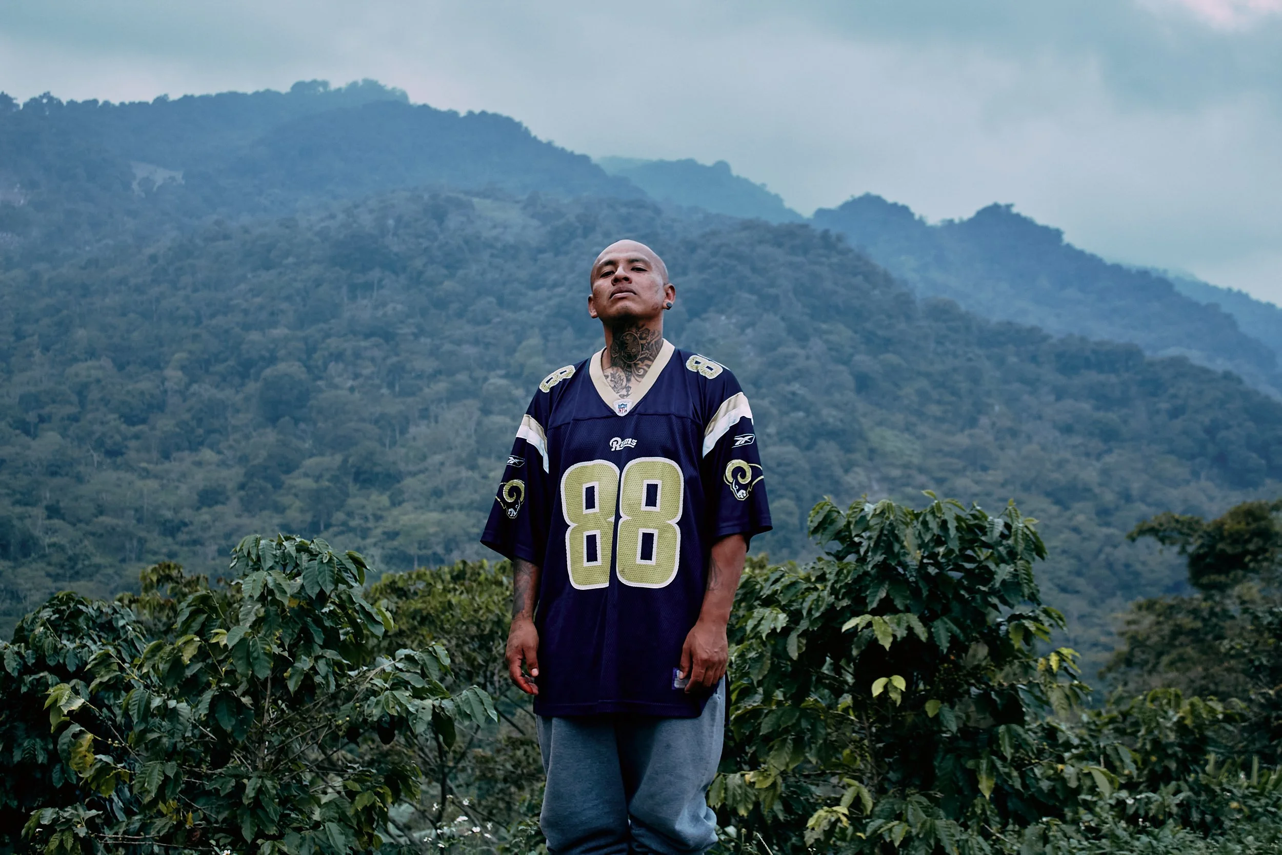 Un hombre con tatuajes en el cuello y cabeza viste una camiseta de fútbol de los Rams de Los Ángeles, de pie en un campo de café con montañas en el fondo.