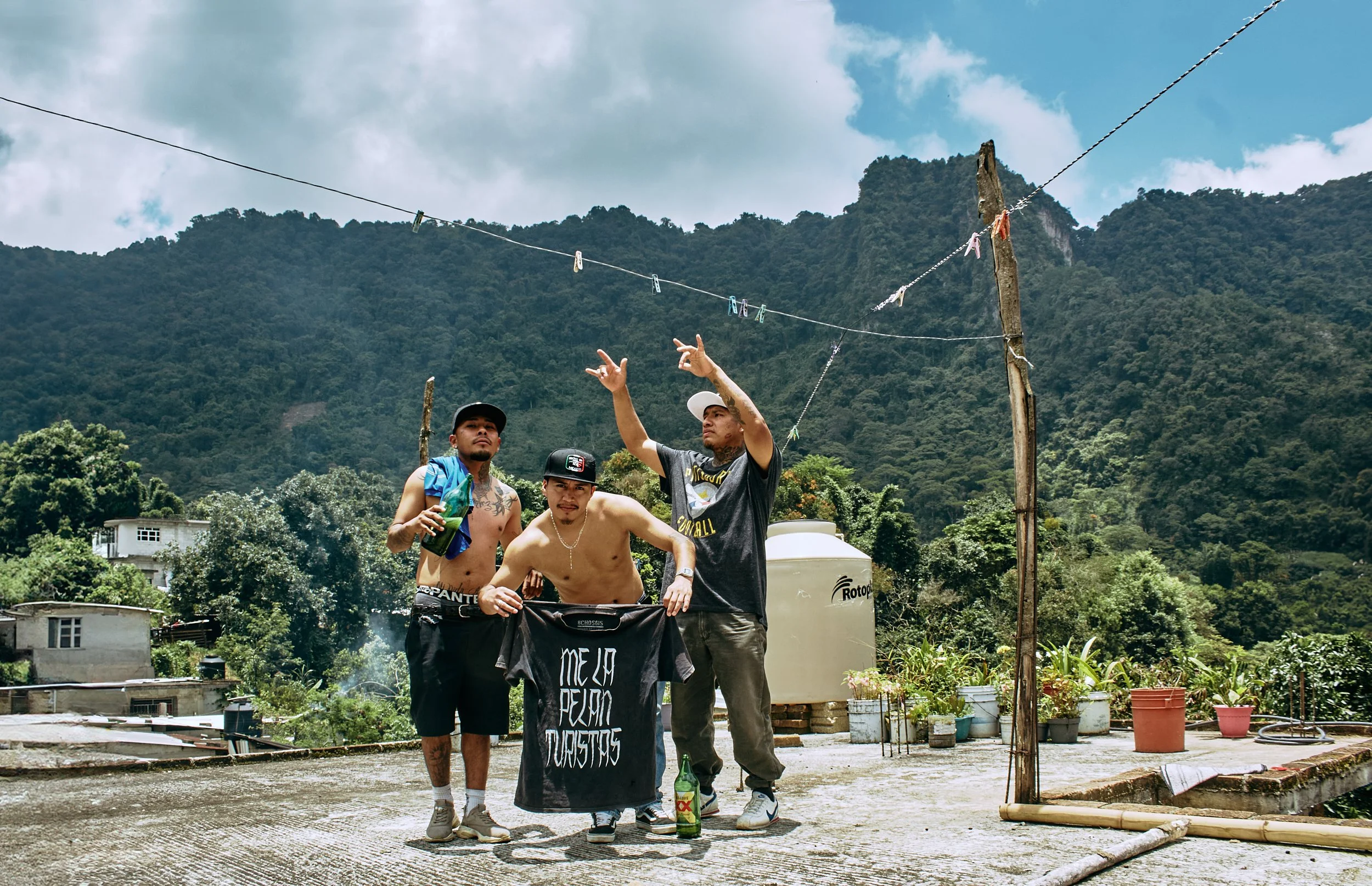 Cuatro jóvenes en un espacio abierto con montañas verdes de fondo, algunos con bebidas y ropa casual, rodeados de utensilios y plantas, parece ser un día de reunión o celebración.